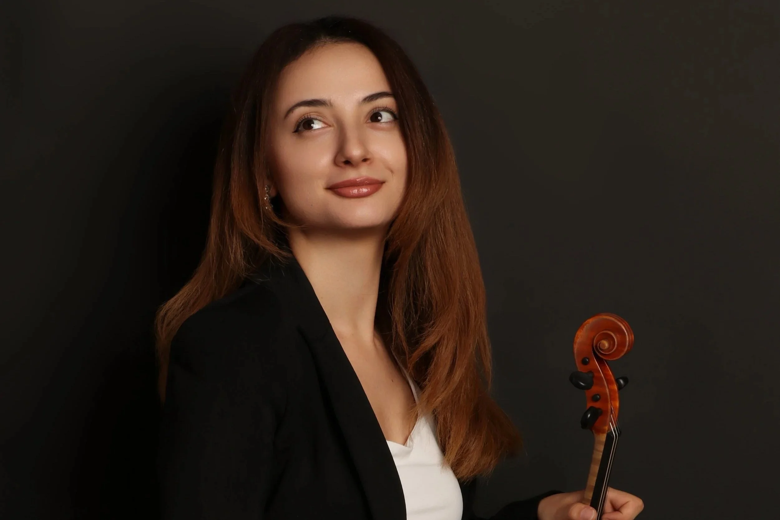 A woman with long brown hair holding a violin against a dark background, wearing a black blazer and a white top.
