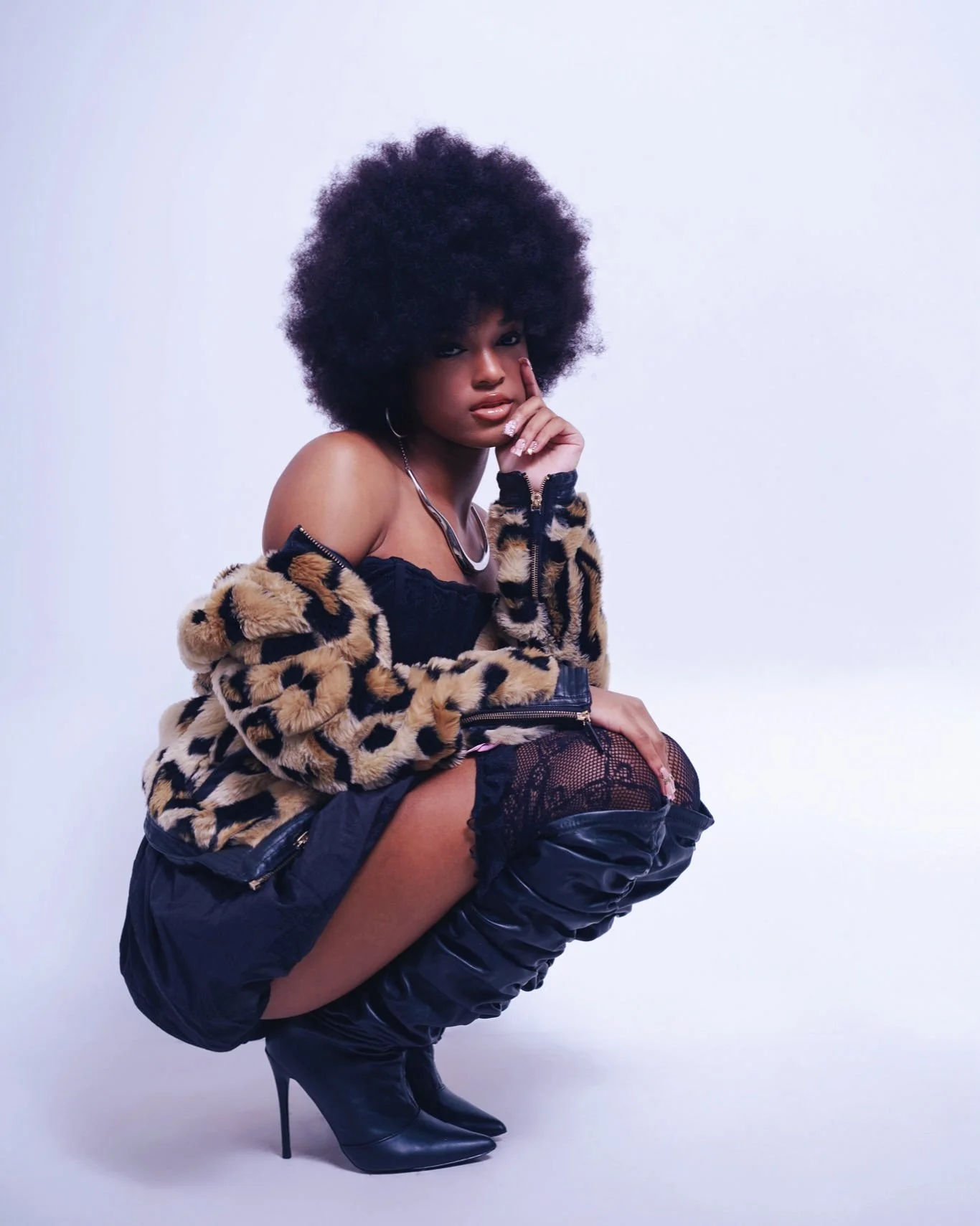 A woman with large curly hair posing with her chin resting on her finger, wearing a leopard print jacket, black lace top, black shorts, fishnet stockings, and thigh-high black boots, against a plain white background.