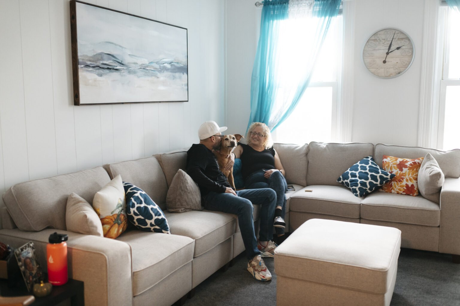 A woman, a man, and a dog sitting on a beige sectional sofa in a living room, with colorful pillows and a large window with blue curtains behind them.