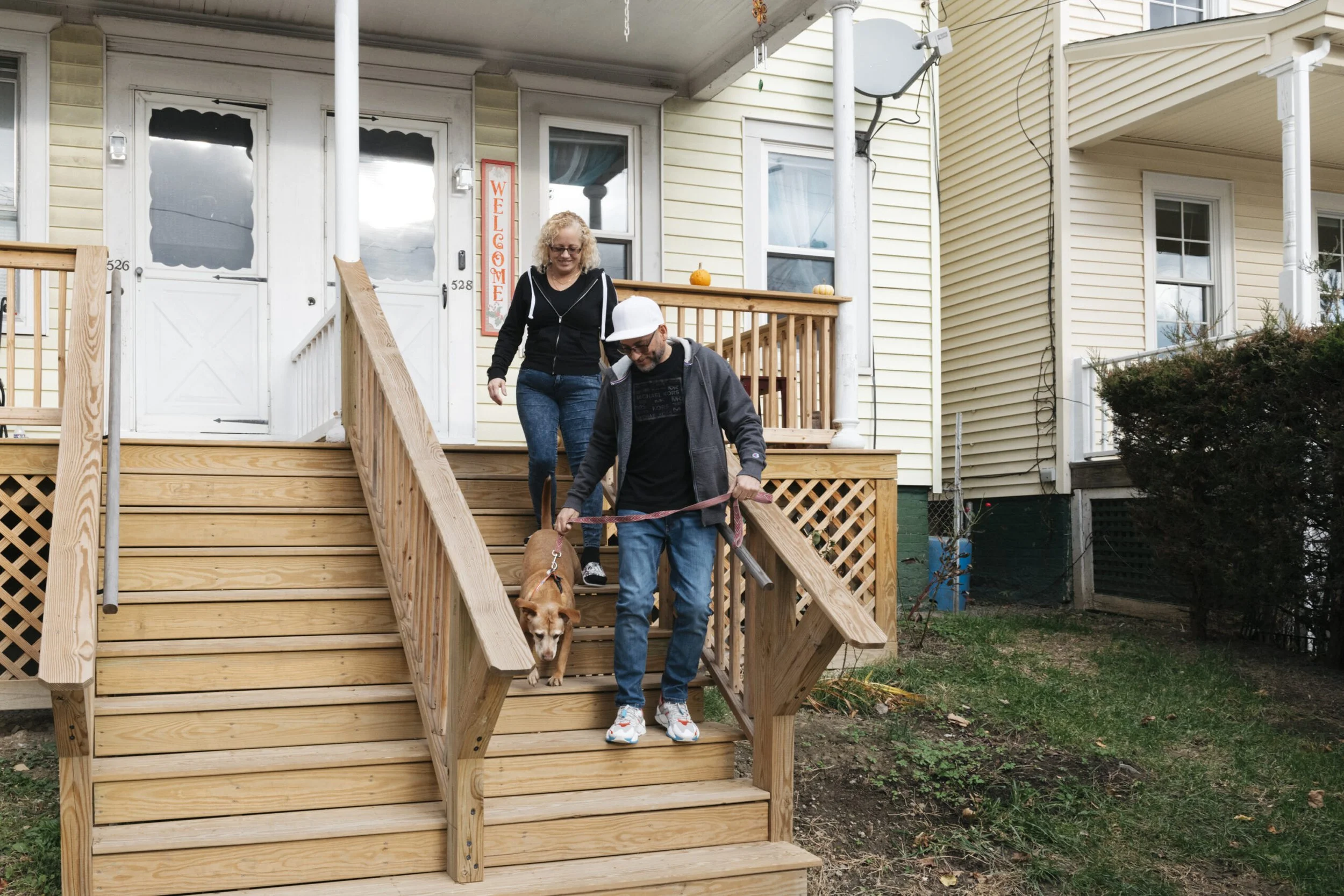 A woman and man walking down outdoor wooden stairs with a dog. The woman has curly blonde hair and is wearing glasses, a black hoodie, and jeans. The man is wearing a white cap, black hoodie, gray jacket, and jeans. The dog is a brown and white corgi on a leash. The background shows a yellow house with white trim, a porch with pumpkin decorations, and various windows.