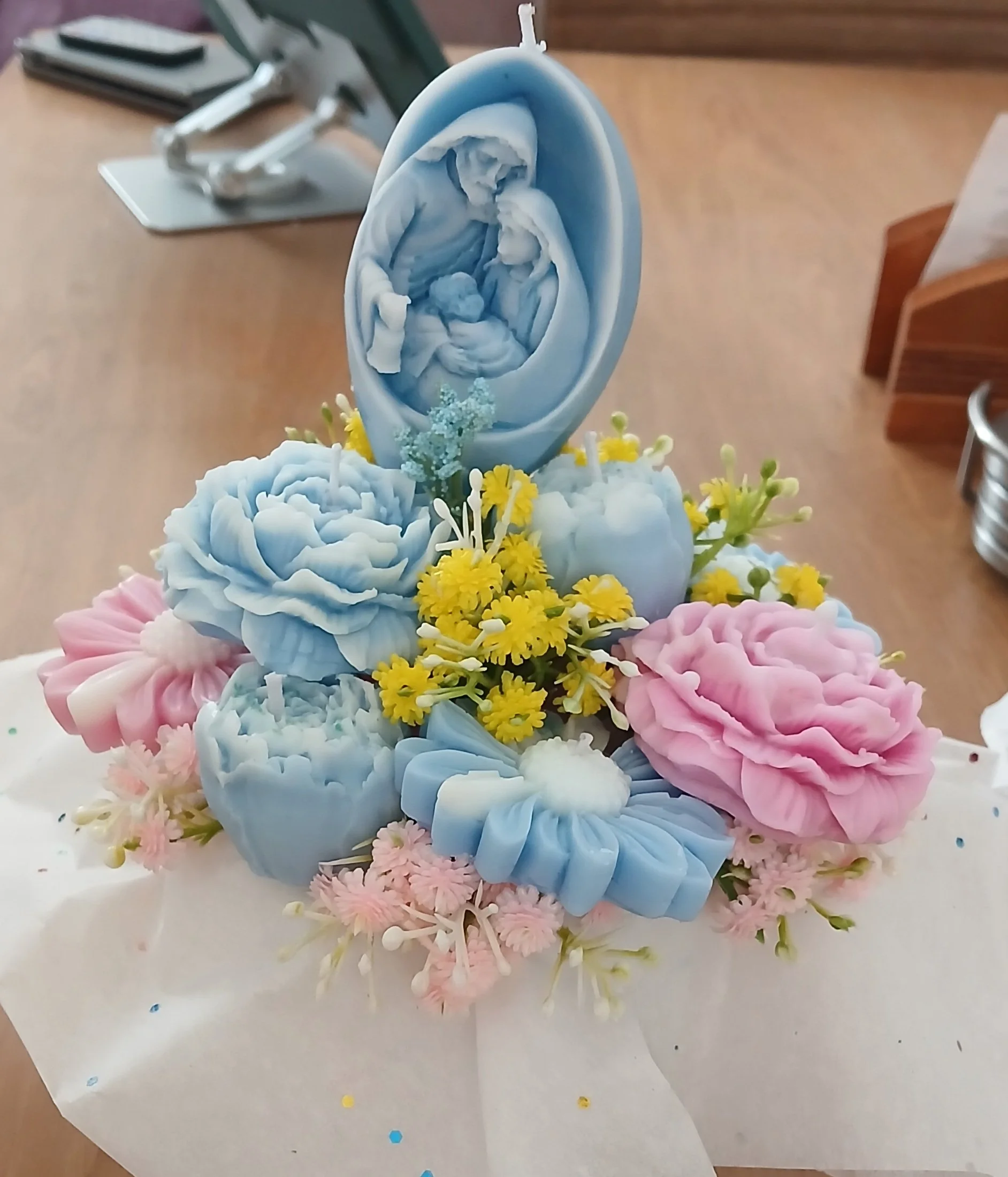 Velas decorativas con figuras religiosas, rodeadas de flores en colores pastel en una mesa, probablemente para una celebración religiosa o especial.