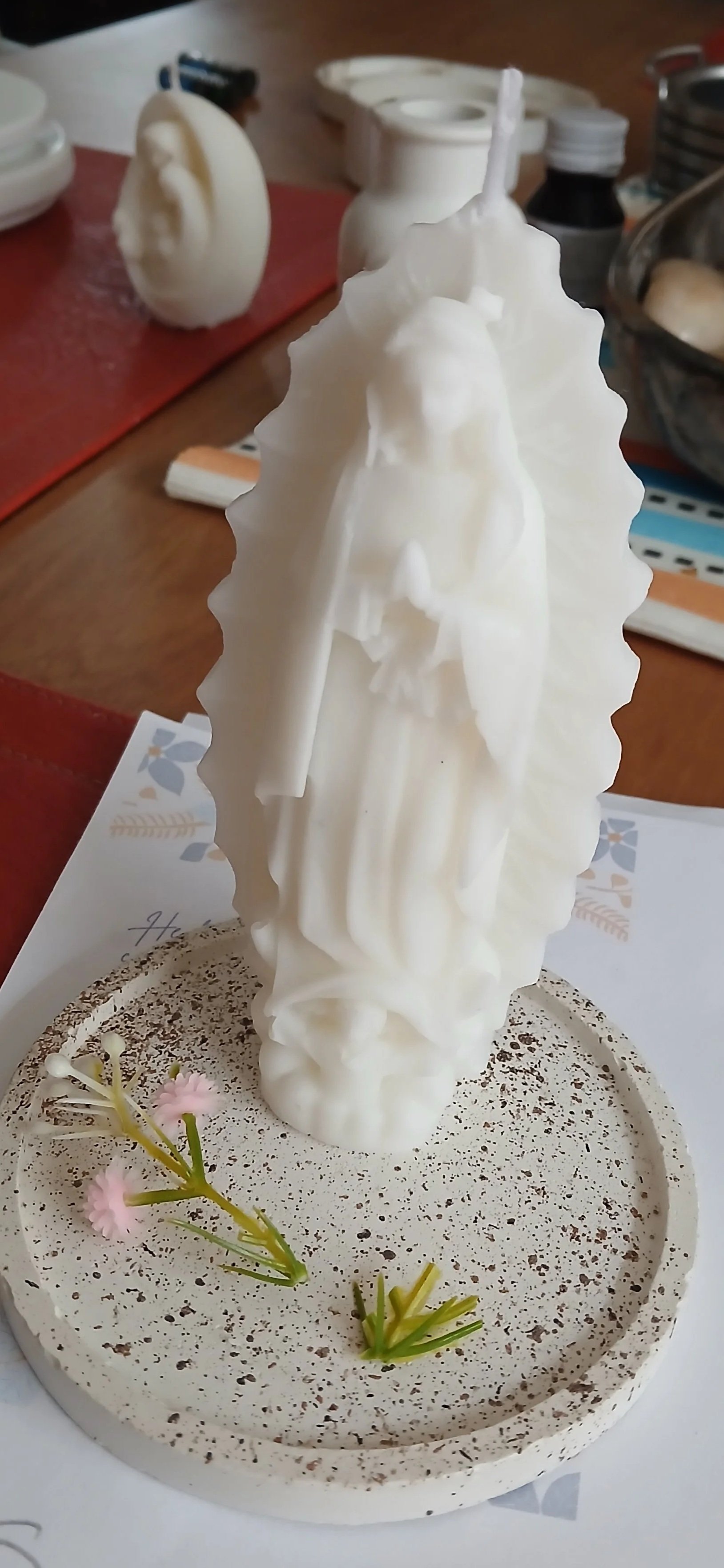 Escultura de color blanco que representa a una figura religiosa, posiblemente la Virgen María, con un niño en sus brazos, colocada en una base decorativa con arena y pequeñas flores artificiales