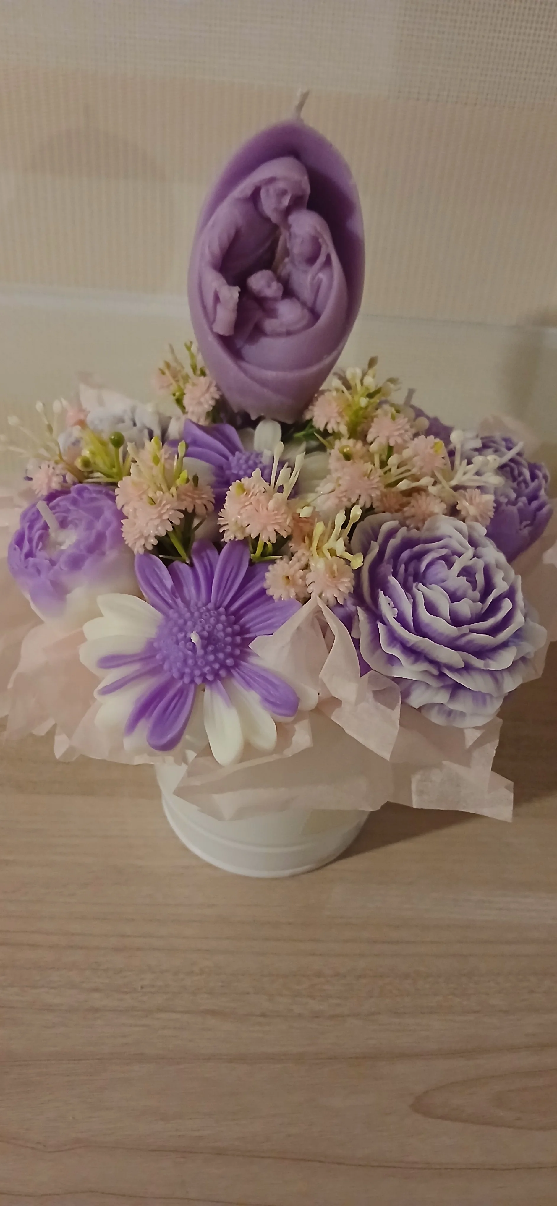 Arreglo floral con flores moradas, blancas y rosadas, incluyendo una figura de madera de la Santa Familia de Nazaret, en una maceta blanca.