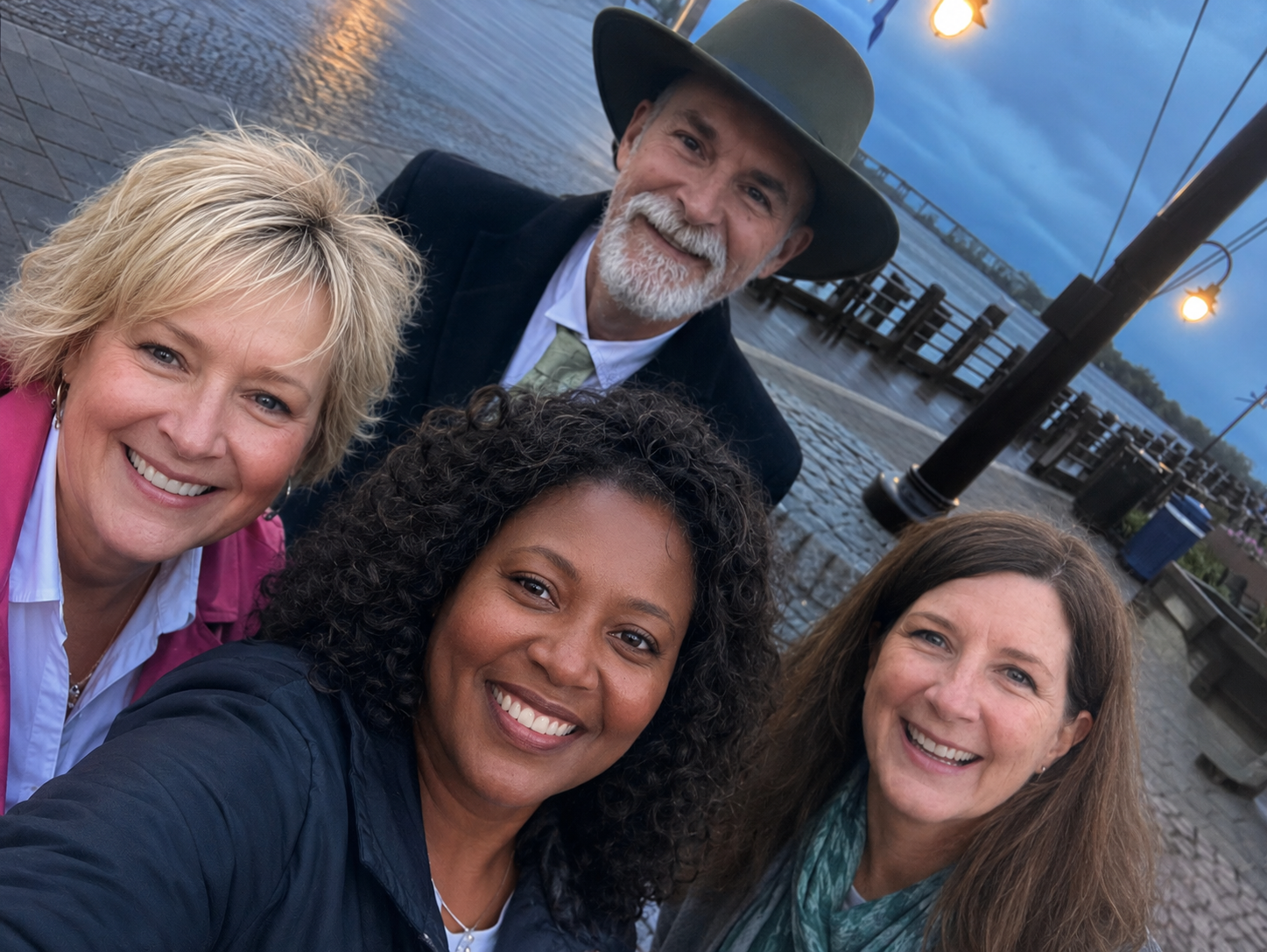 Group of five smiling women and a man taking a selfie near a waterfront during sunset, with a pier and cloudy sky in the background.