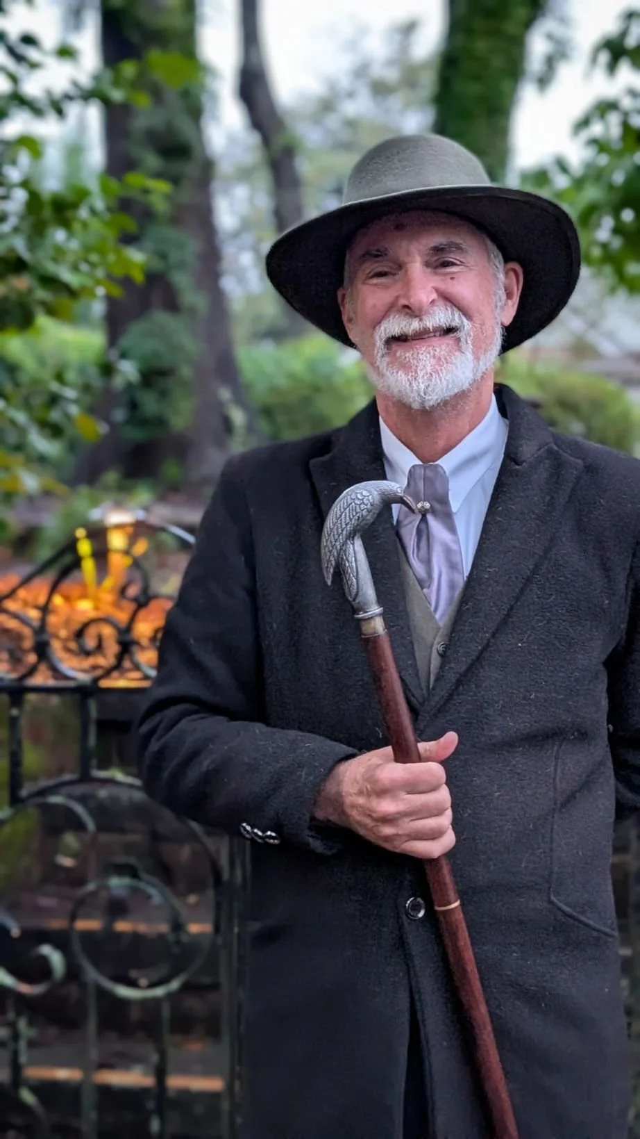 An elderly man with a white beard and mustache, wearing a dark coat and a wide-brimmed hat, stands outdoors holding a cane with a snake-shaped handle. There are trees and a decorative metal fence in the background.