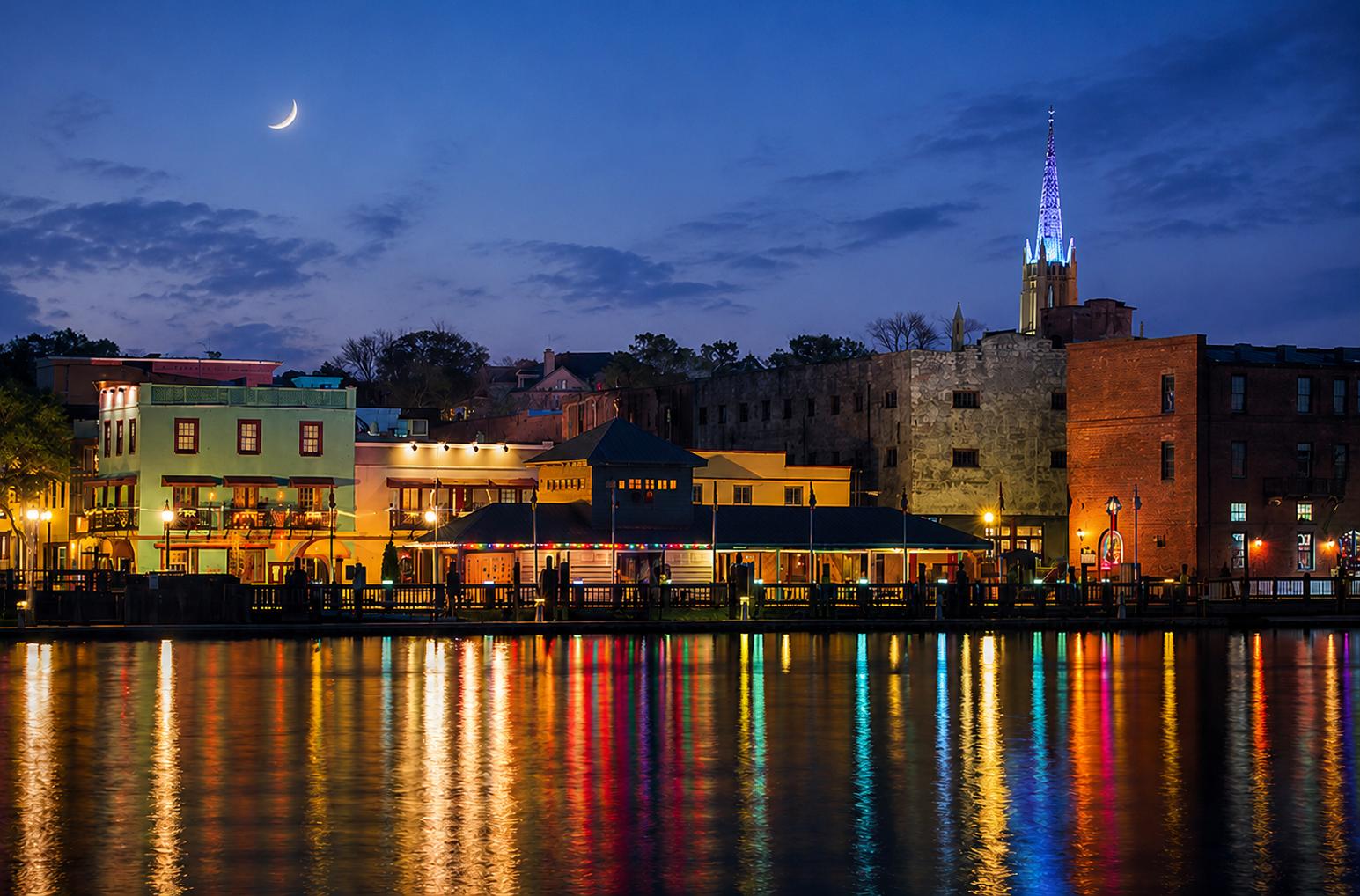 Nighttime view of a colorful waterfront cityscape with a church steeple illuminated in blue in the background, reflections on the water, and a crescent moon in a partly cloudy sky.