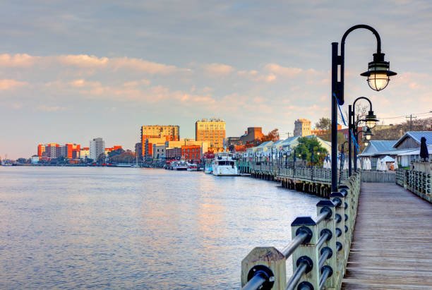 View of a river with buildings in the background, a dock with lamps, and a boat docked along the shoreline.
