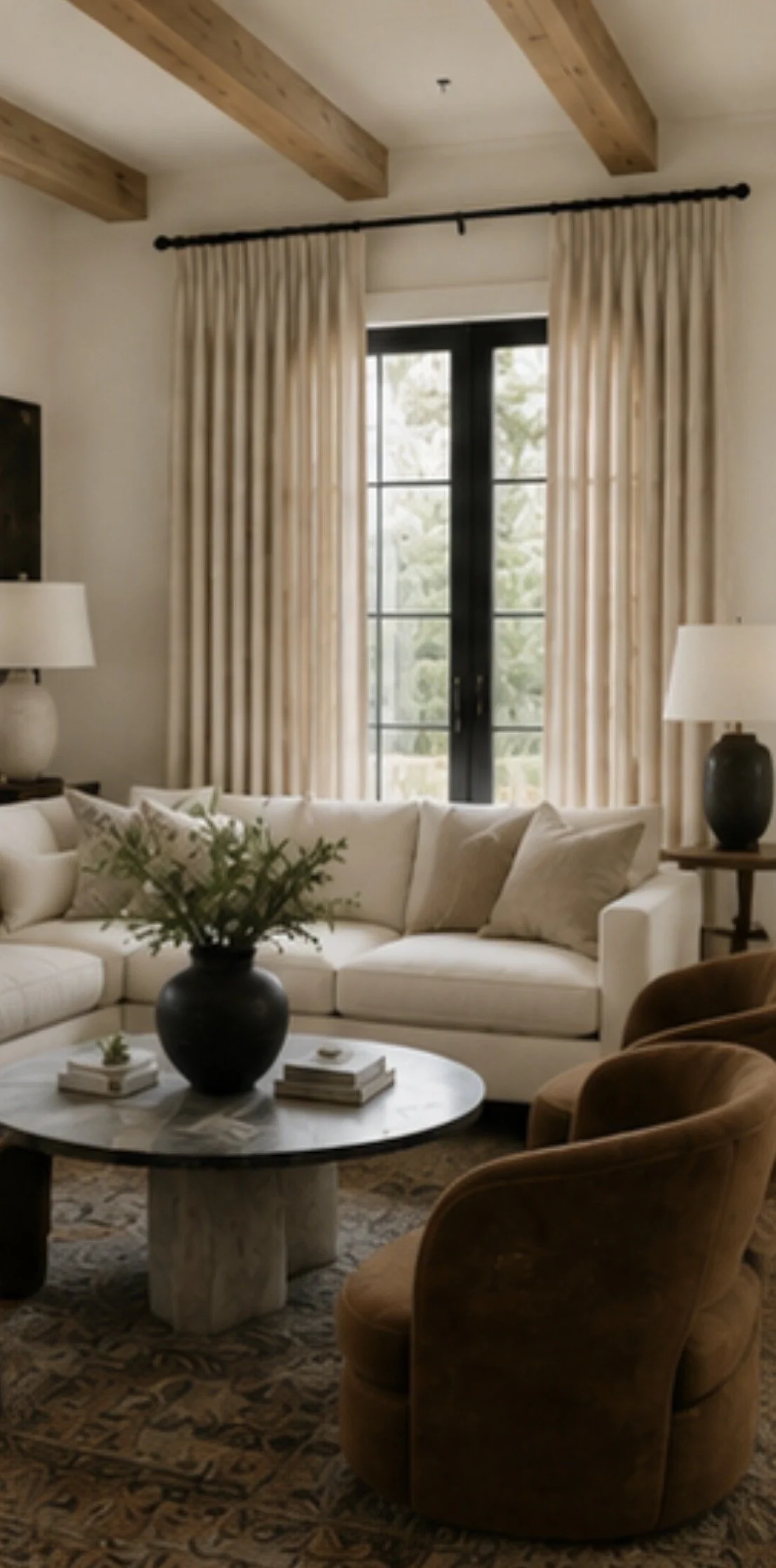 Living room with beige curtains, a white sofa with pillows, a black vase with greenery on a round marble coffee table, a brown armchair, and side tables with lamps, large windows, and wooden ceiling beams.