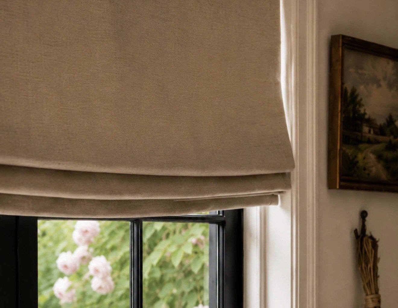 Close-up of beige Roman window shade partially pulled down, covering part of a black-framed window with green and pink flowering plants outside; framed landscape painting on wall to the right.