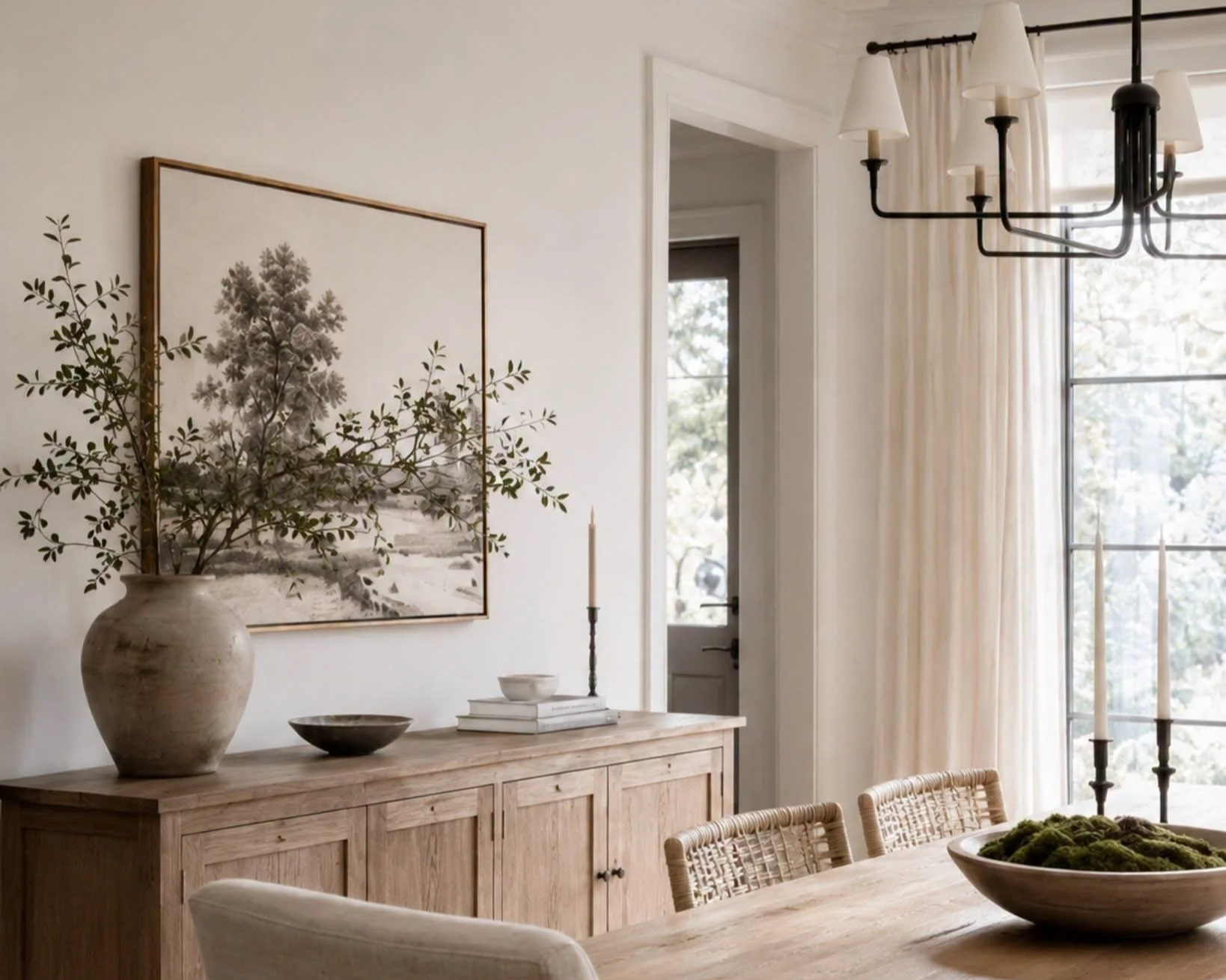 A dining room with a wooden sideboard decorated with a large vase filled with greenery, a candle, and bowls. A white framed black and white tree print hangs on the wall, and there are tall candles on the table. The room has large windows with curtains and a black chandelier.