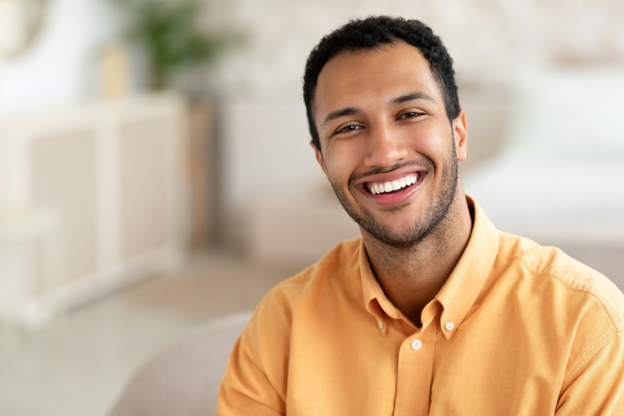 A young man with short curly dark hair and a beard is smiling while looking at the camera, wearing a yellow button-up shirt, in a bright indoor setting with a blurred background.