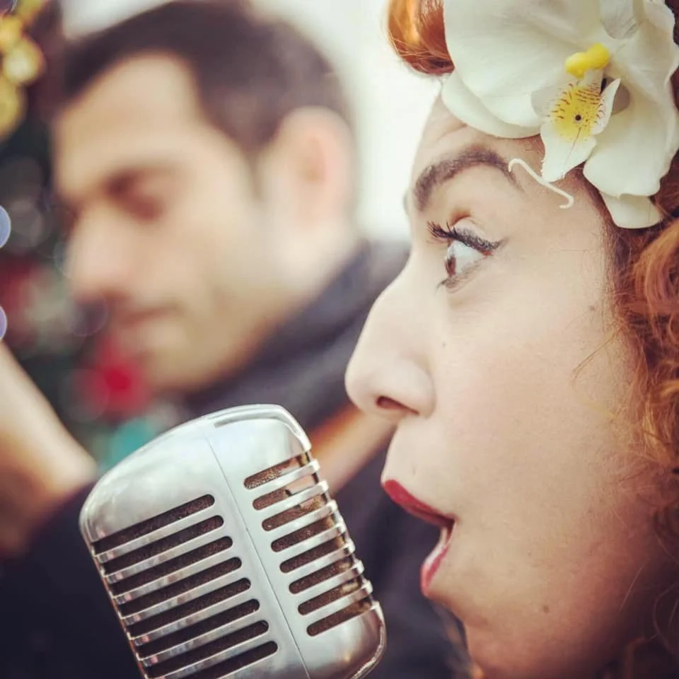 A woman with curly red hair, wearing a flower headpiece, is singing into a vintage microphone with a man in the background.