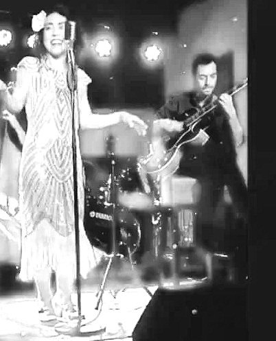 A woman singing on stage with a microphone, flanked by a bassist and a guitarist, under red stage lights at a live music performance.