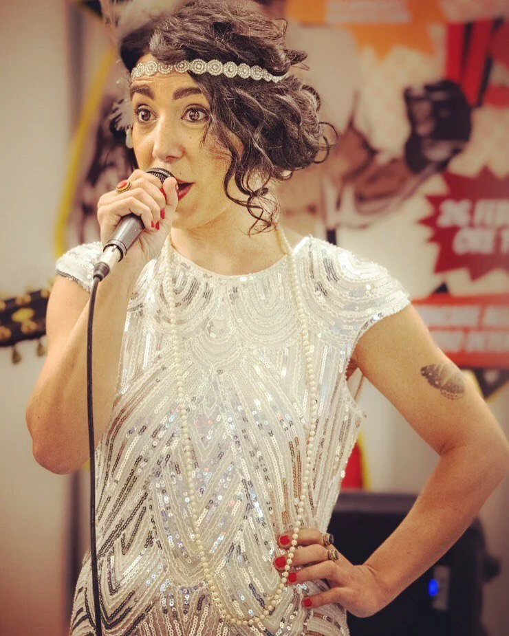 A woman with curly hair wearing a headband, a sparkly vintage-style dress, and holding a microphone, appears to be performing or speaking at an event.