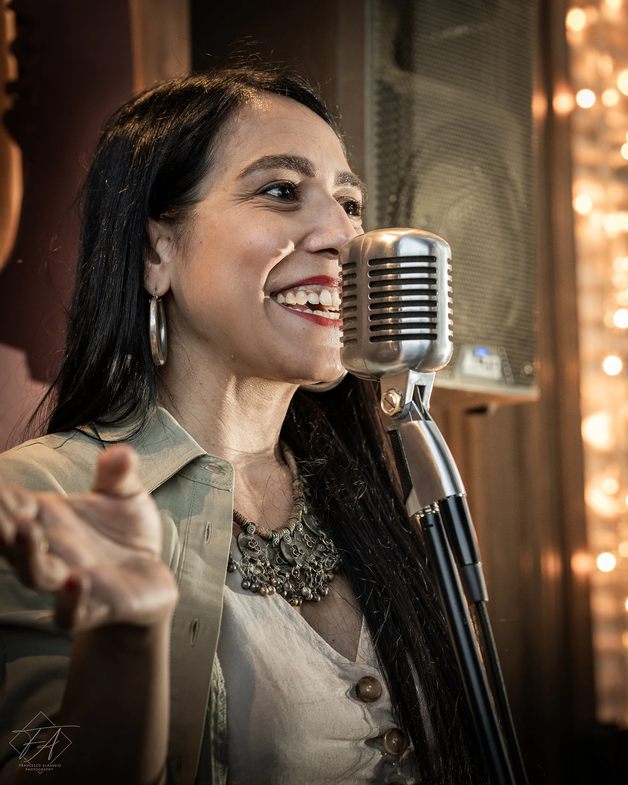 A woman with long dark hair, smiling, speaking into a vintage microphone, wearing earrings, a necklace, and a beige top, in a warmly lit indoor setting.