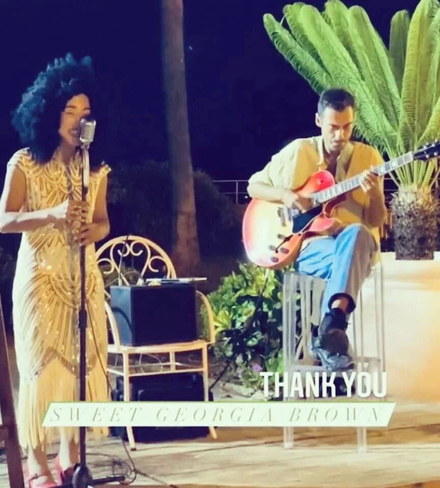 A woman singing into a vintage microphone and a man playing an electric guitar outdoors at night, with large green plants behind them. The image includes a thank you message and the name Georgia Brown.