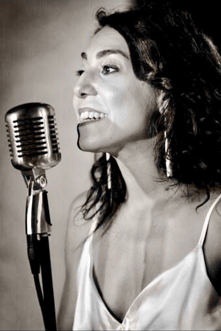 A woman singing into a vintage microphone, smiling with her hair styled in loose waves, in a black and white photograph.