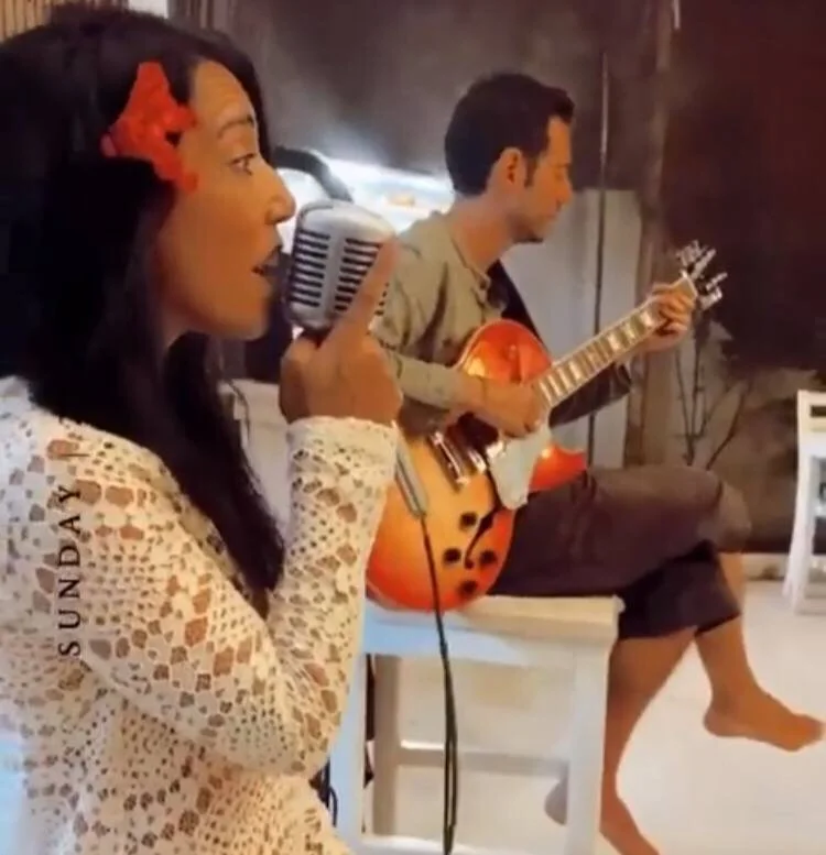 A woman singing into a vintage microphone with a red flower in her hair, sitting next to a man playing an orange acoustic guitar. They are indoors, sitting on a white bench or chair.