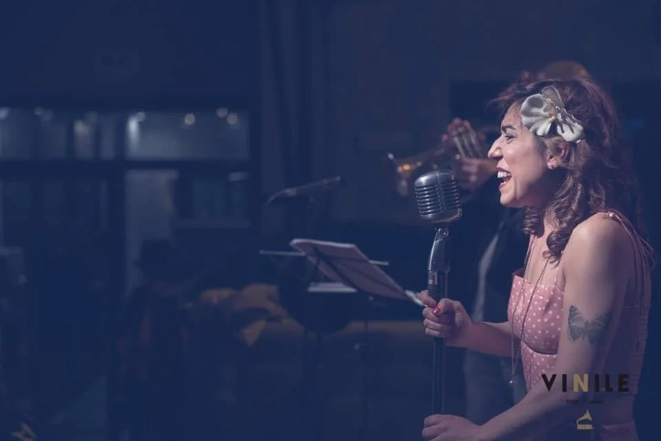 A woman singing into a vintage microphone with a band playing in the background.