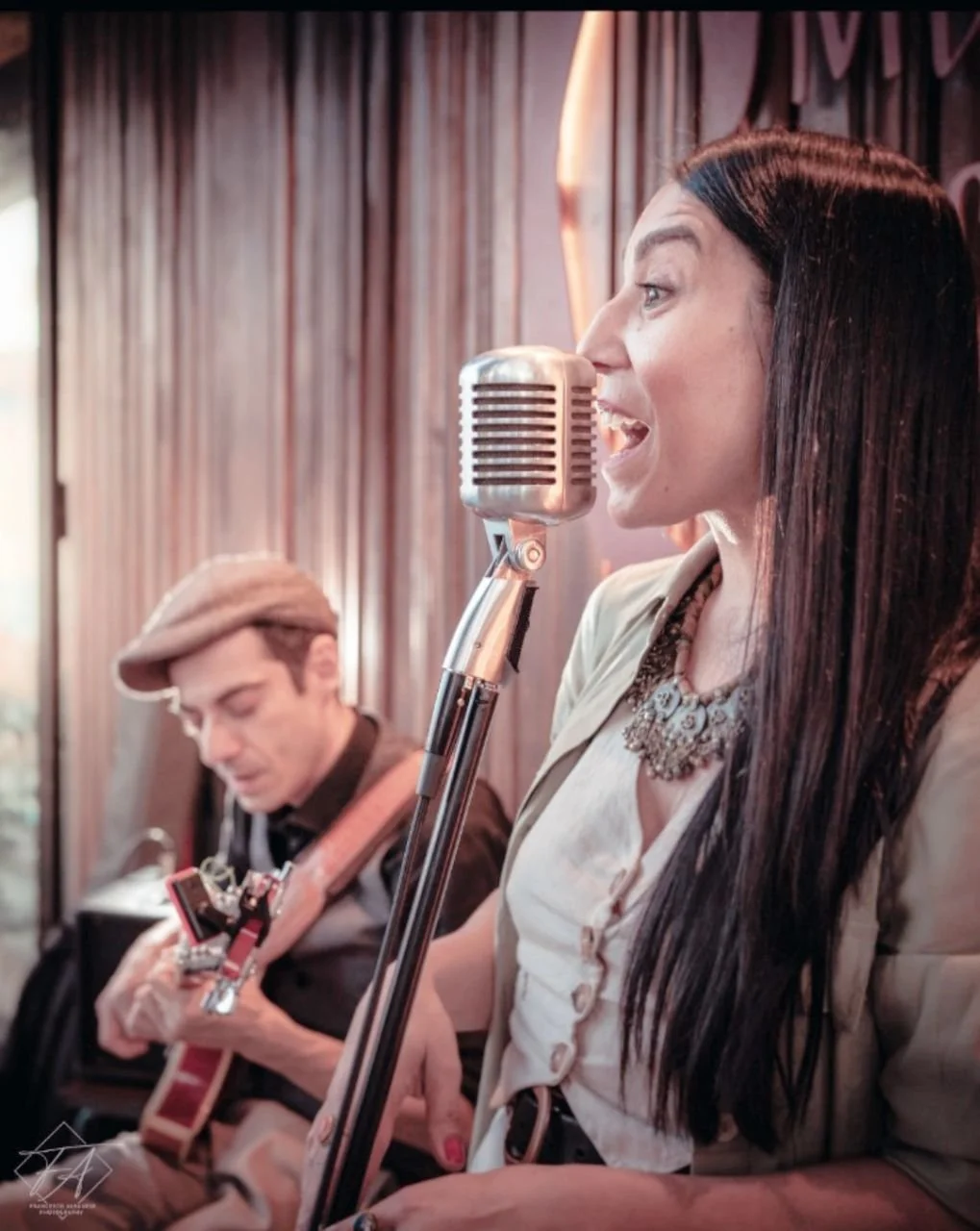 A woman singing into a vintage microphone while a man plays guitar in the background at a cozy indoor venue.
