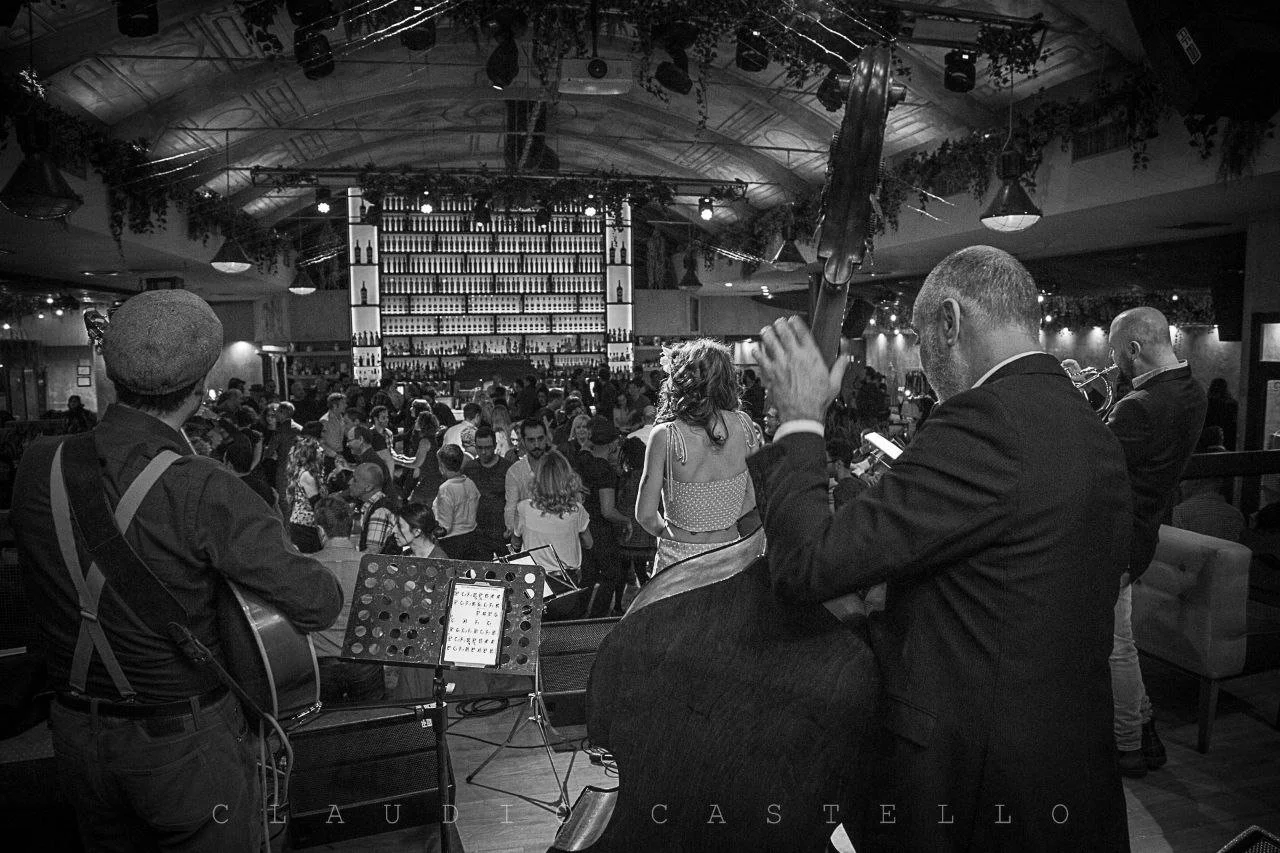 Jazz band performing on stage with audience in a bar or jazz club with bottles on shelves in the background