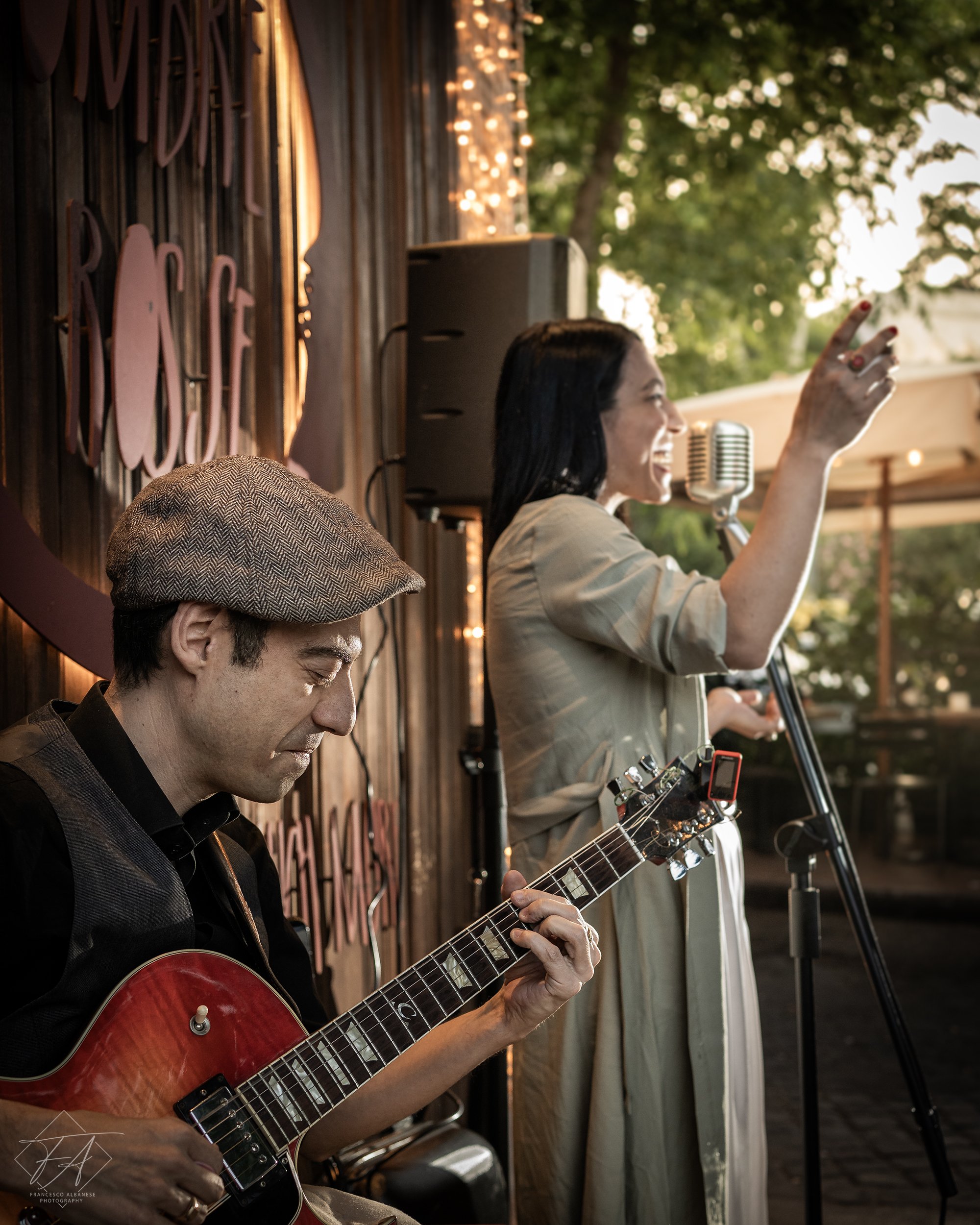 Musicians performing outdoors at sunset, one playing guitar and another singing into a microphone, with string lights and trees in the background.