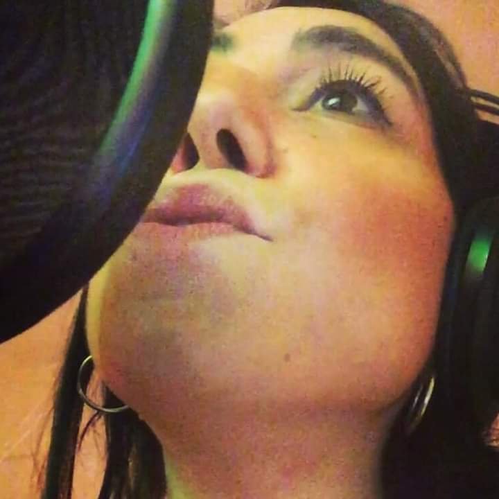 A woman with dark hair and hoop earrings singing into a microphone with headphones on.