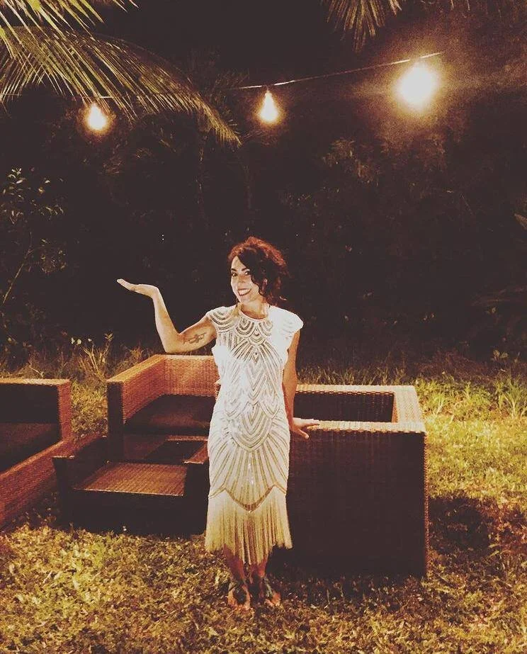 A woman with short dark hair, wearing a white and gold dress, stands outdoors at night with string lights overhead. She is smiling, with one arm extended and the other behind her, in front of wicker furniture and greenery.