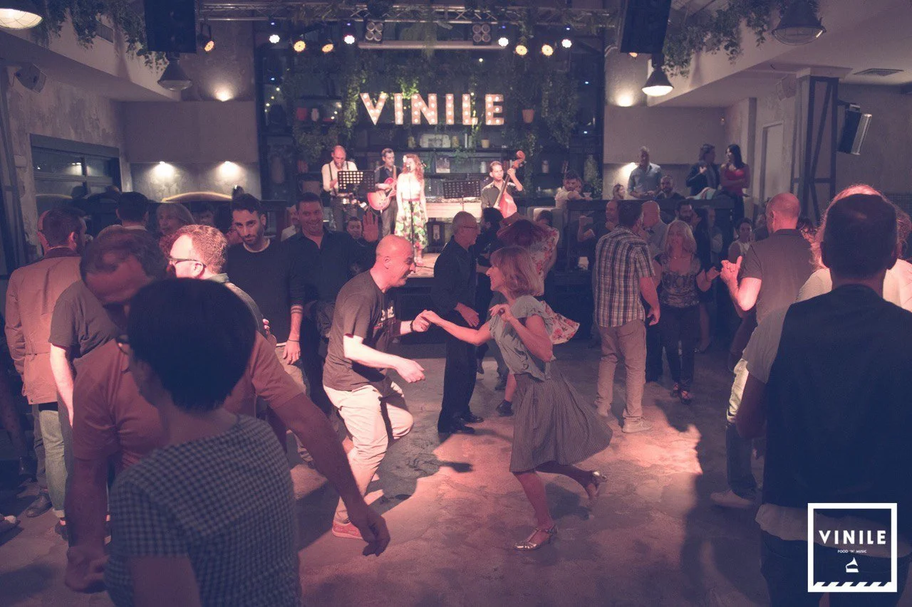 People dancing and socializing in a dimly lit venue with a live band playing on stage in the background, decorated with greenery and the word 'VILNE' illuminated above the stage.