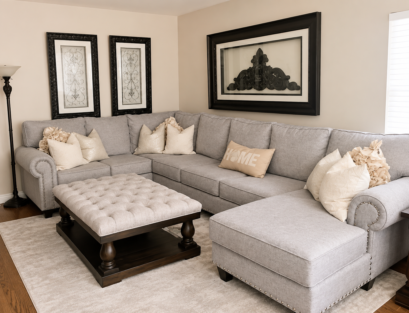 A living room with a light gray sectional sofa, decorative pillows, a tufted ottoman, and framed artwork on the walls.