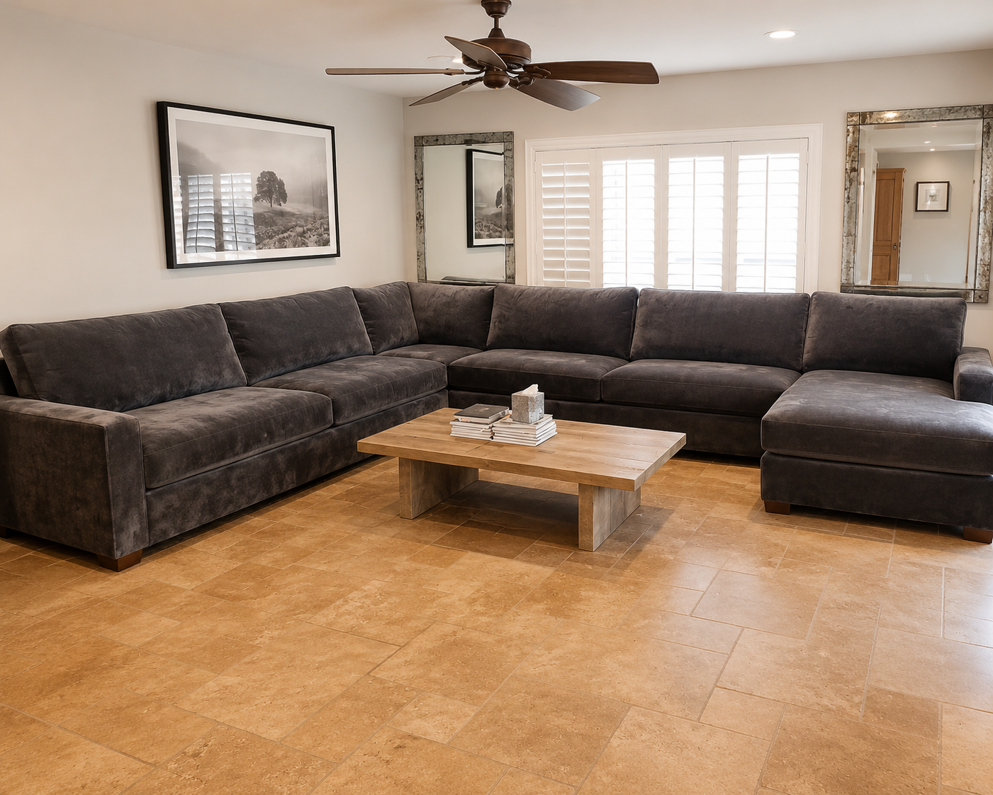 Living room with a large dark gray sectional sofa, a wooden coffee table with books and tissue box, and two framed black-and-white landscape photographs on the wall. There are three large mirrors, a window with white shutters, a ceiling fan, and wood