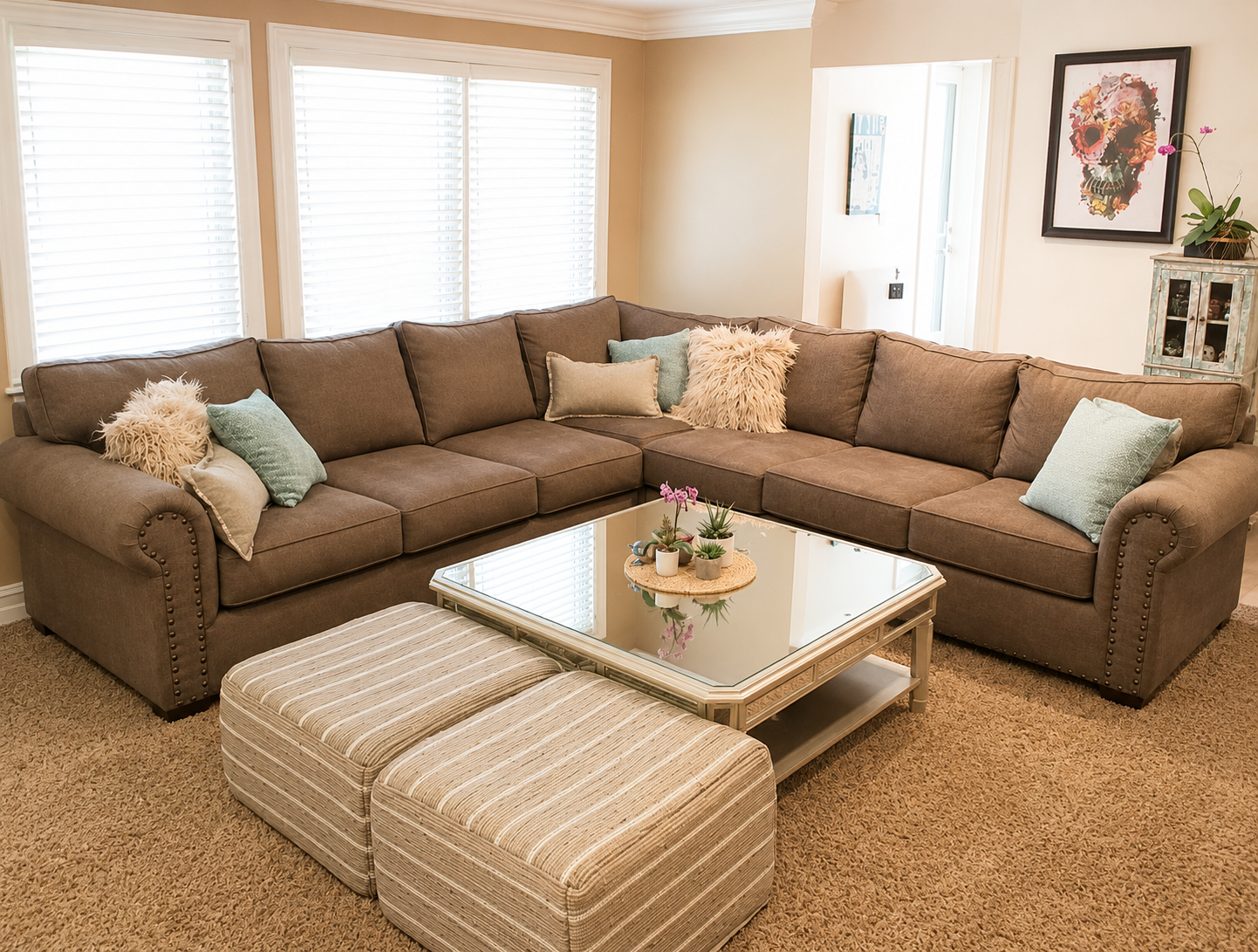 Living room with a large L-shaped brown sofa decorated with colorful pillows, a glass-topped coffee table with potted plants, two striped ottomans, window blinds, and wall art.