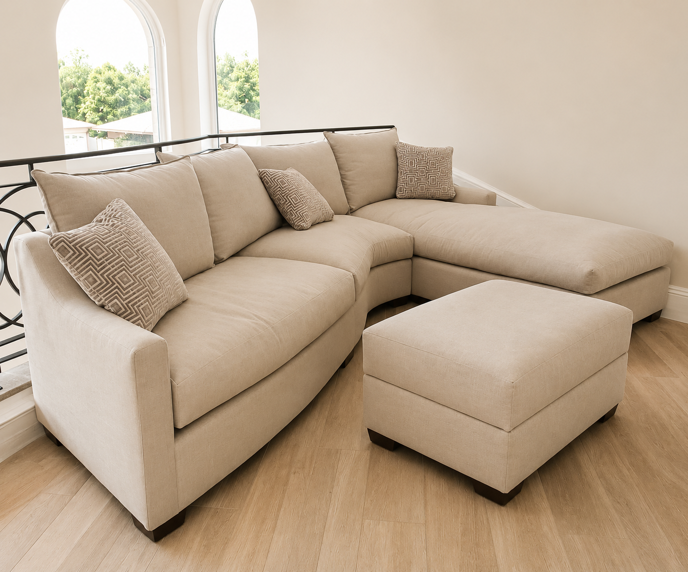 Light beige sectional sofa with four throw pillows, and a matching ottoman in a room with light wood flooring, large arched windows, and a view of trees outside.
