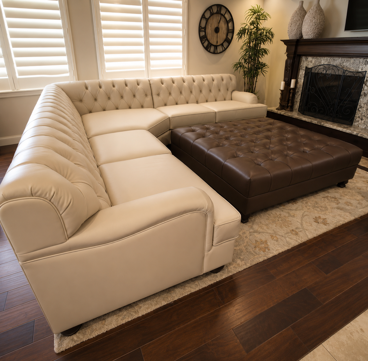 Living room with a large beige L-shaped tufted leather sofa, brown tufted ottoman, dark wooden fireplace mantel, decorative vases, a large round wall clock, green indoor plant, and plantation shutters on windows.