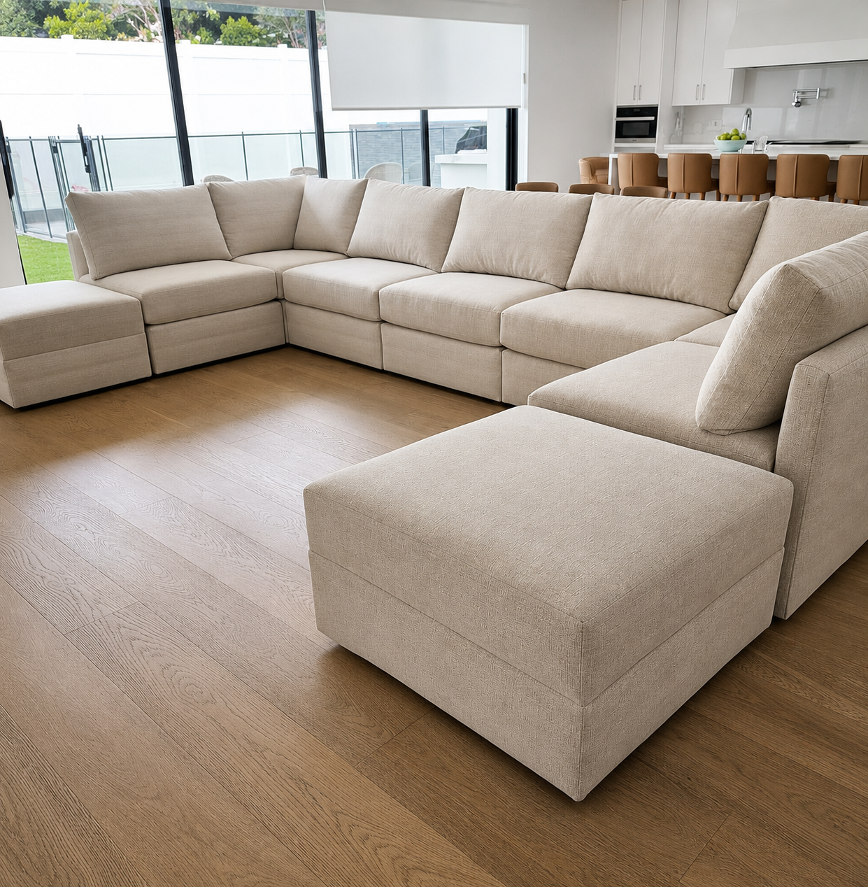 A spacious beige sectional sofa with matching ottoman in a modern living room with wooden flooring and large windows.