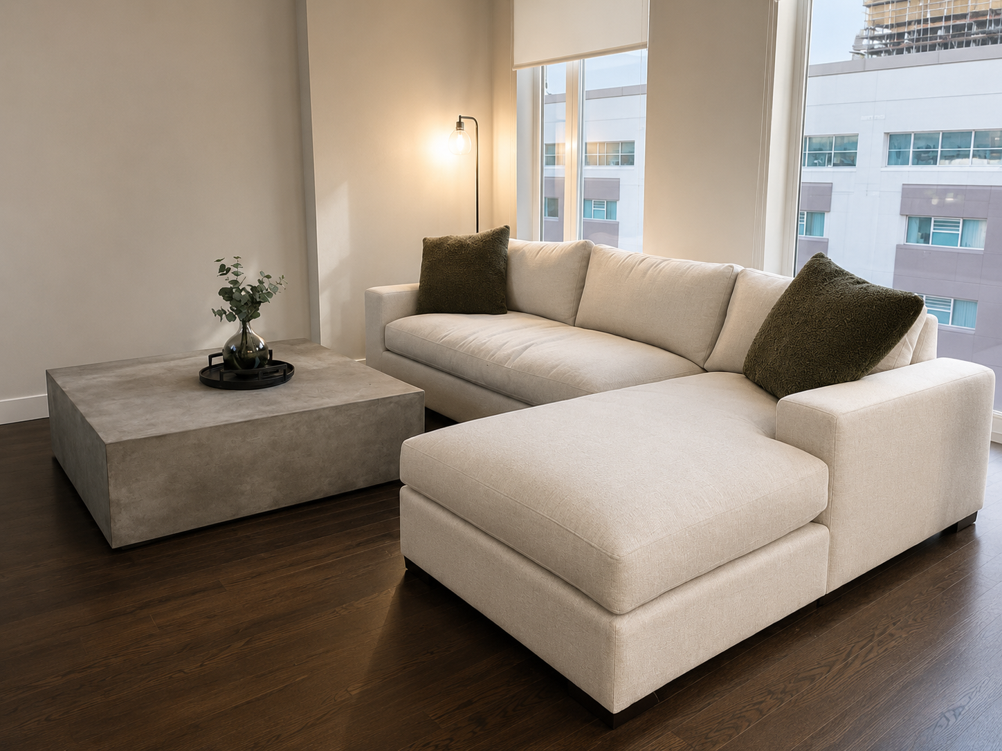 Modern living room with beige sectional sofa, dark green throw pillows, concrete coffee table with vase of greenery, hardwood floors, and large windows showing apartment buildings outside.