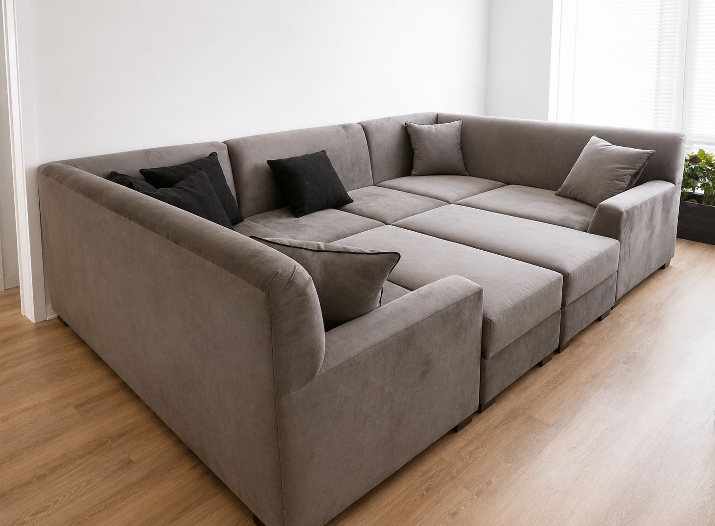 A large L-shaped gray sectional sofa with five pillows, three black and two gray, in a bright living room with light wooden flooring and white walls.