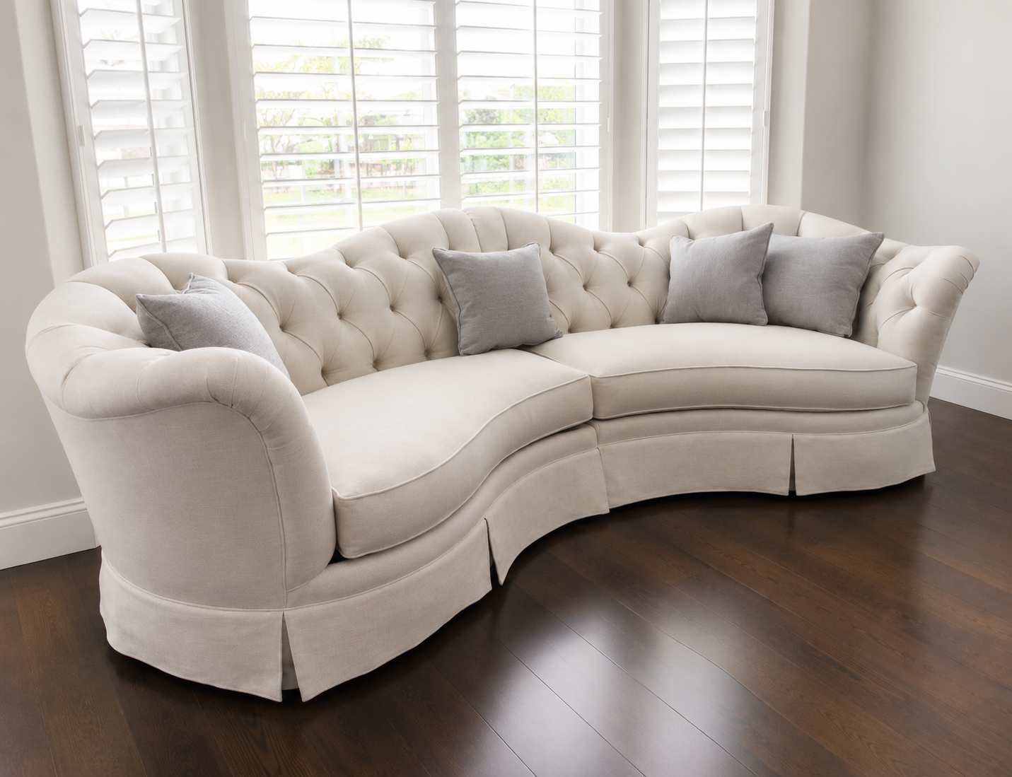 A cream-colored tufted sofa with rolled arms and a pleated skirt, decorated with four gray throw pillows, in front of large windows with white plantation shutters, dark hardwood floors.