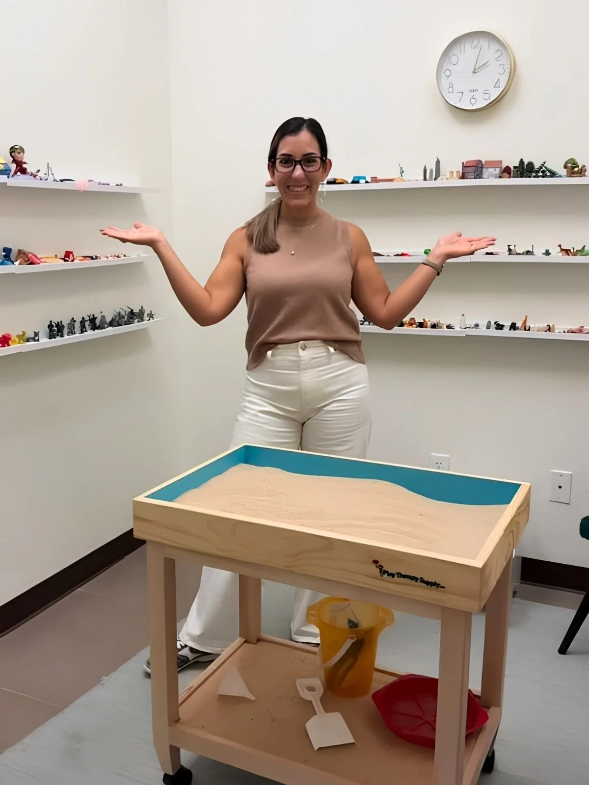 Dr. Clarissa Salinas standing beside a sandtray therapy table surrounded by shelves of miniature figurines at Heartsease Counseling Center.