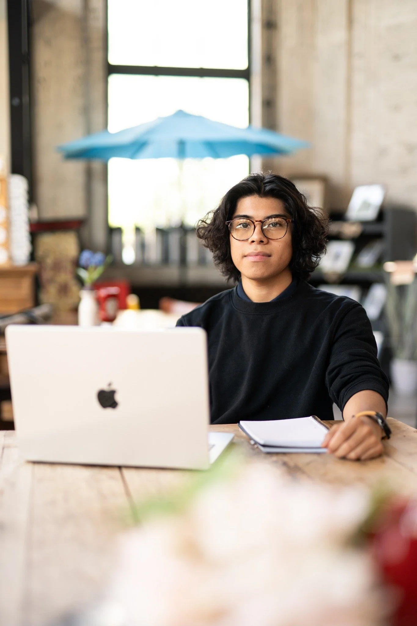 A young woman with curly dark hair and glasses sitting at a wooden table in a cozy cafe or workspace, with an open laptop and a notebook in front of her, looking directly at the camera.