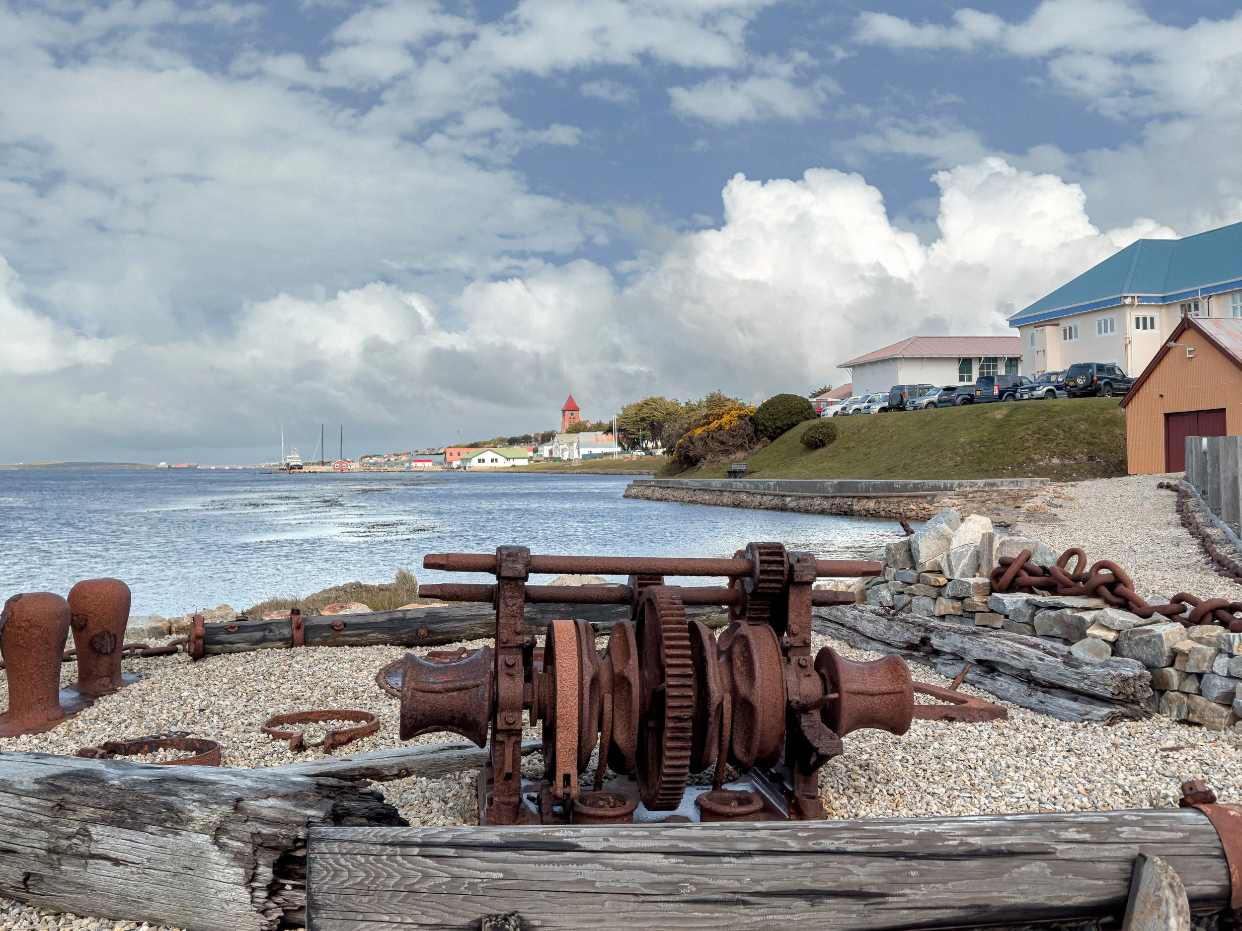 A waterfront scene with boats in the water, buildings with colorful roofs, and a partly cloudy sky, with rusted ship parts and chains in the foreground.