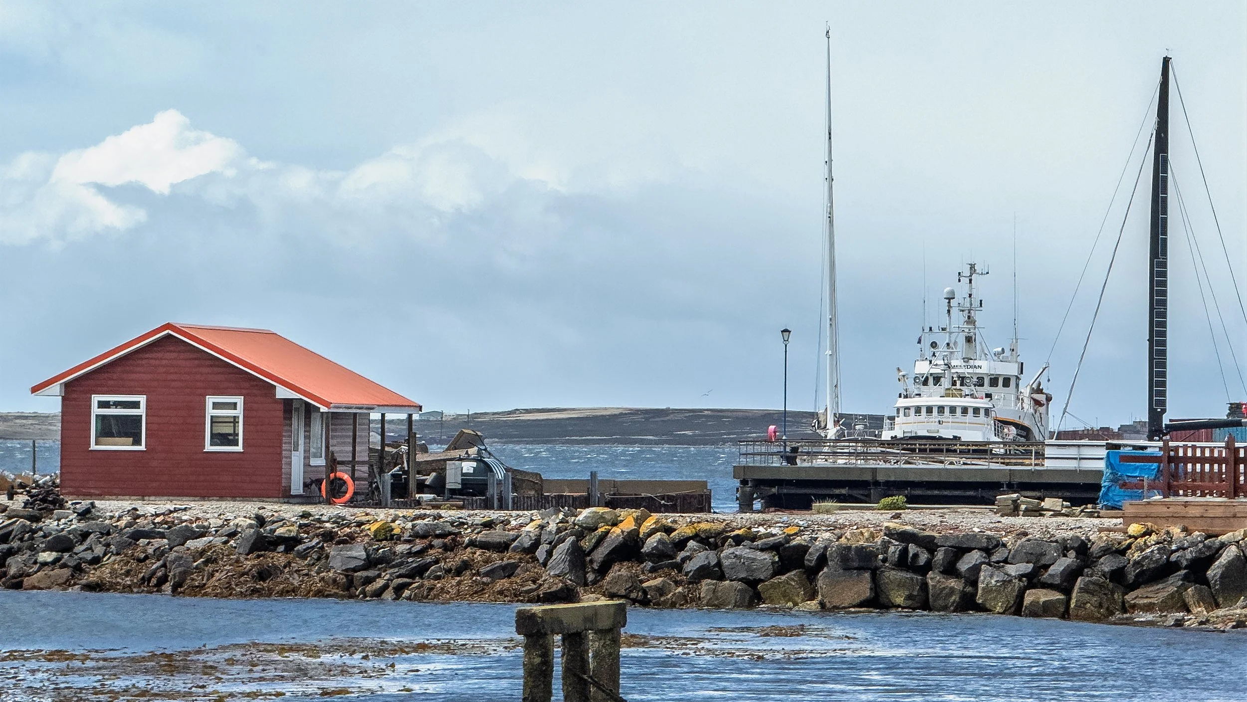 A scenic harbor scene featuring a red wooden building with a metal roof, a boat docked at the pier, and a lighthouse in the background under a partly cloudy sky.