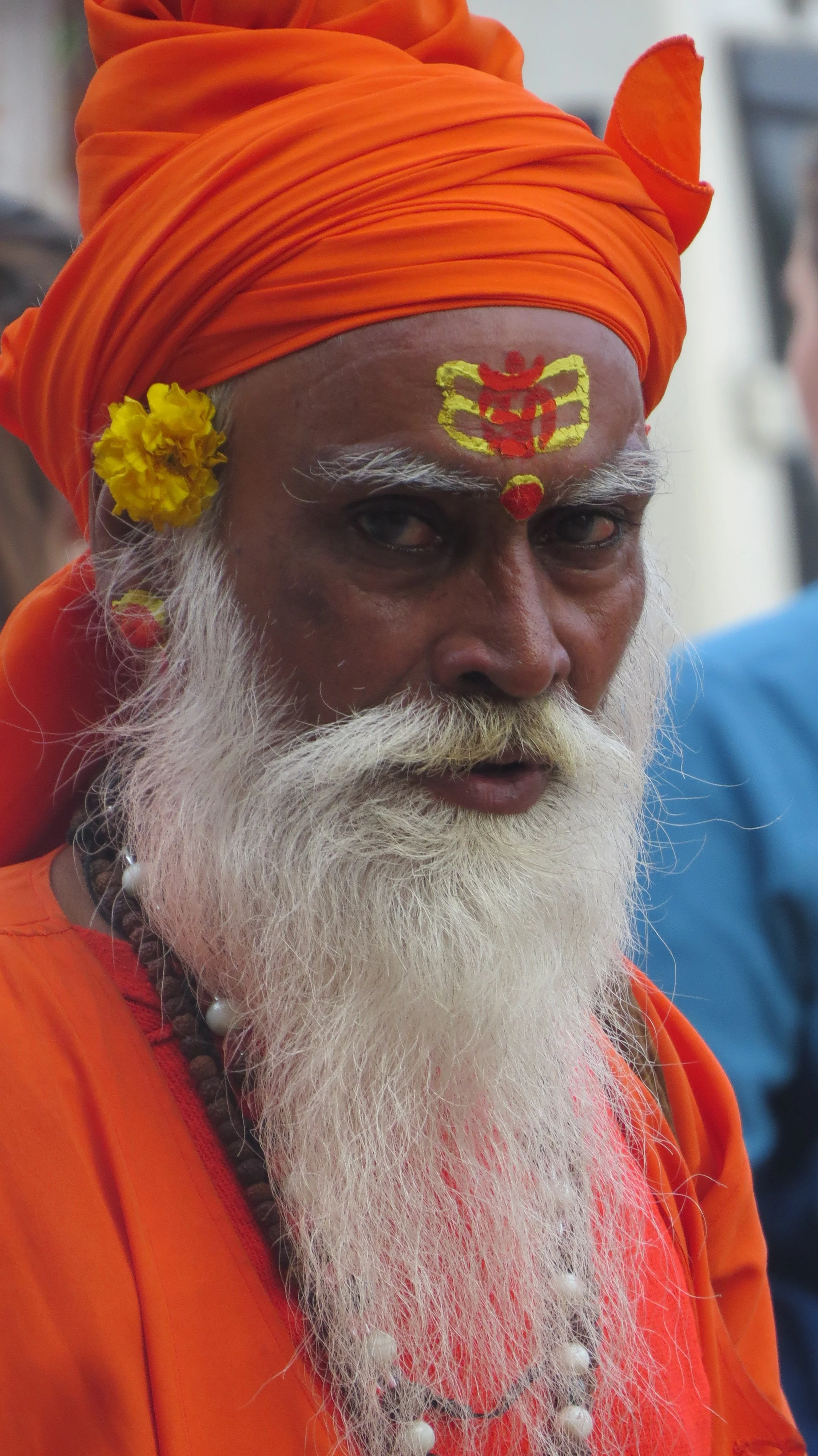 A man with a long white beard and mustache, wearing an orange turban and orange clothing. He has hand-painted red and yellow marks on his forehead, a yellow flower tucked behind his ear, and is wearing a beaded necklace.