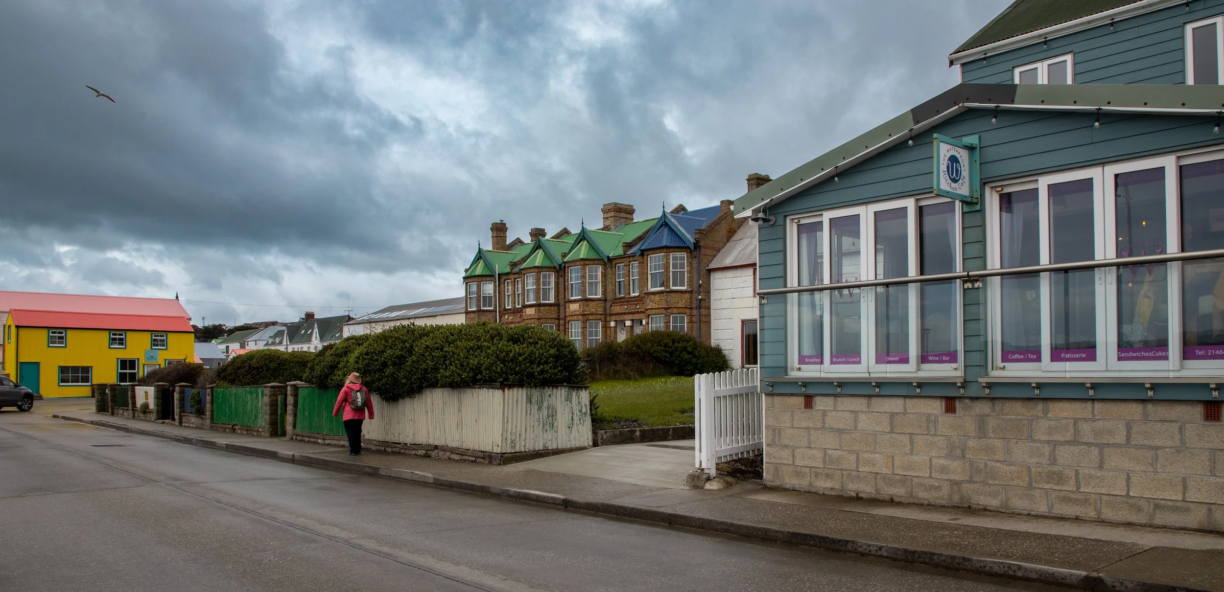 A street scene with colorful buildings under a cloudy sky, including a blue building with large windows and signs for a cafe, a yellow building, and a row of houses with green and blue roofs. A person wearing a pink jacket and backpack walks along th