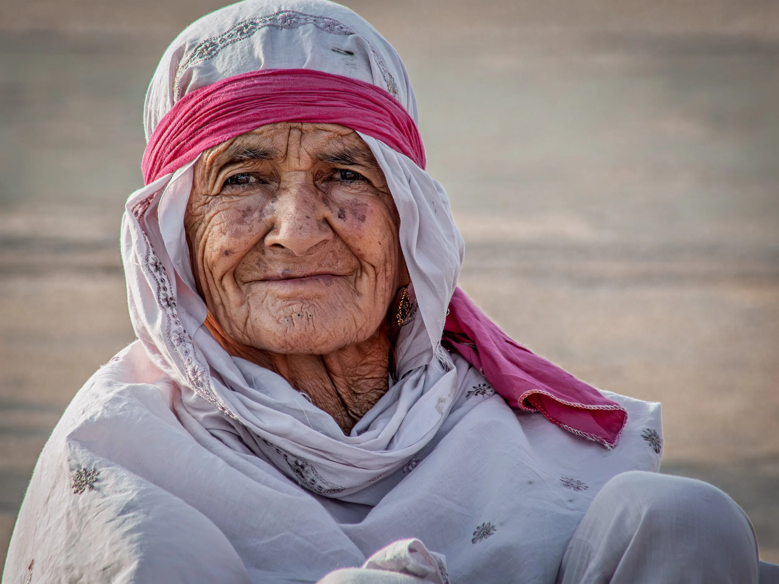 Close-up of an elderly Saudi woman with dark skin and deep wrinkles, wearing a white headscarf with embroidery and a pink scarf around her head, sitting outdoors on a sandy ground.