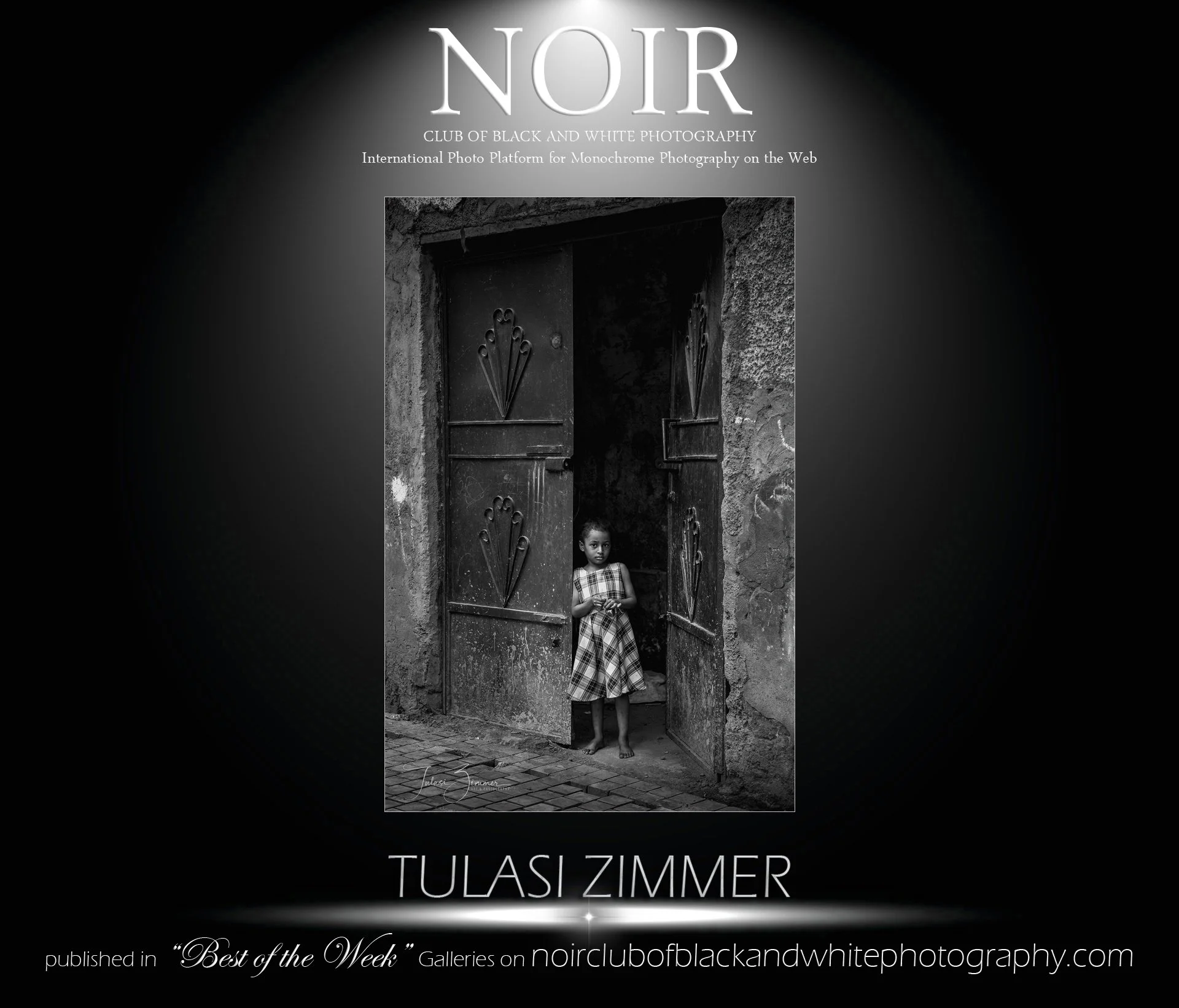 Black and white photograph of a young girl standing in a doorway of a rustic building. The girl is wearing a plaid dress and is barefoot. The doorway has decorative metalwork on the doors and a rough stone wall around it. The photograph has a somber,