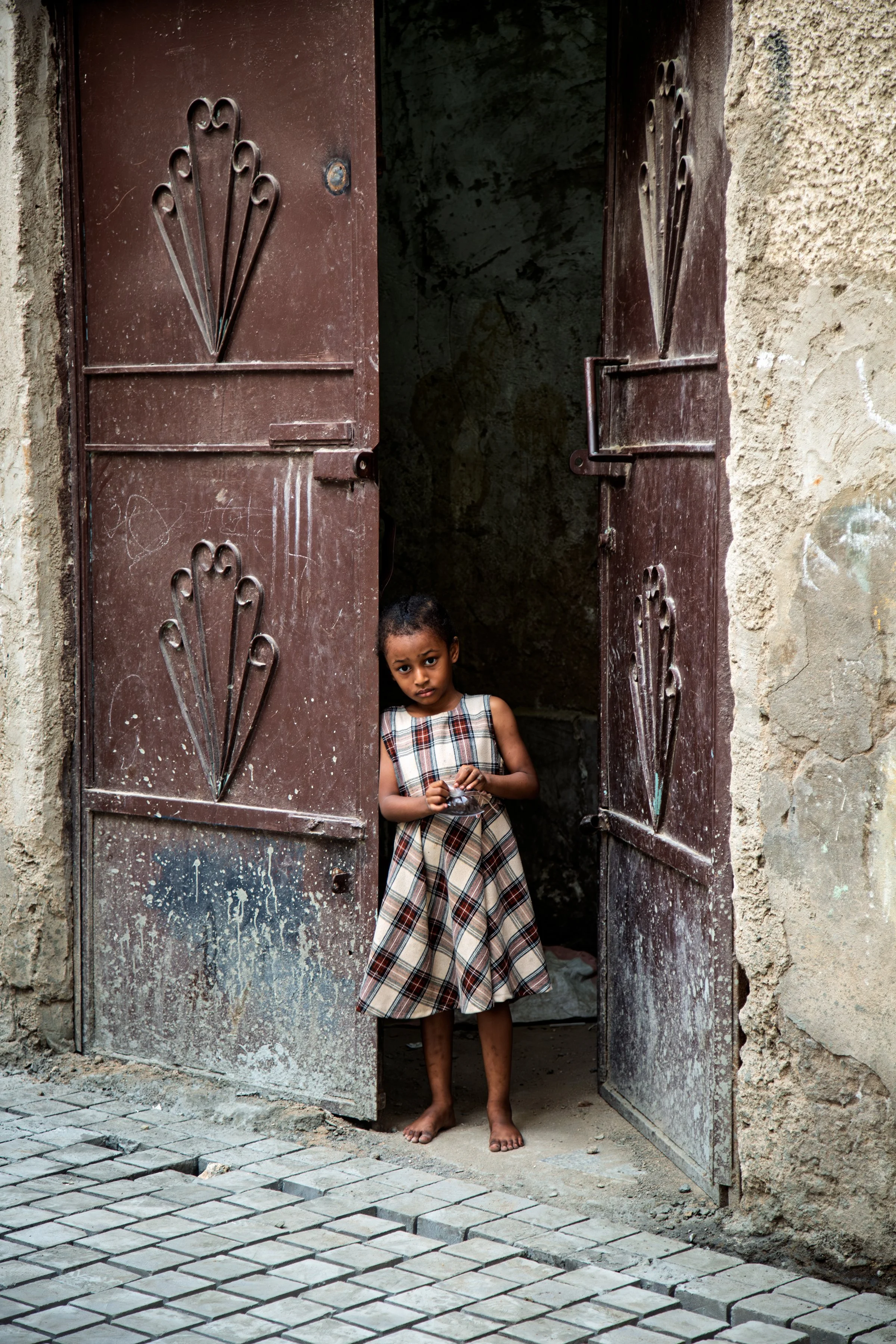 A young girl standing in a doorway with weathered metal double doors. The girl is barefoot, wearing a plaid dress, and appears to be looking at the camera. The surroundings show rough, unpainted wall surfaces and a partially paved street in front of 