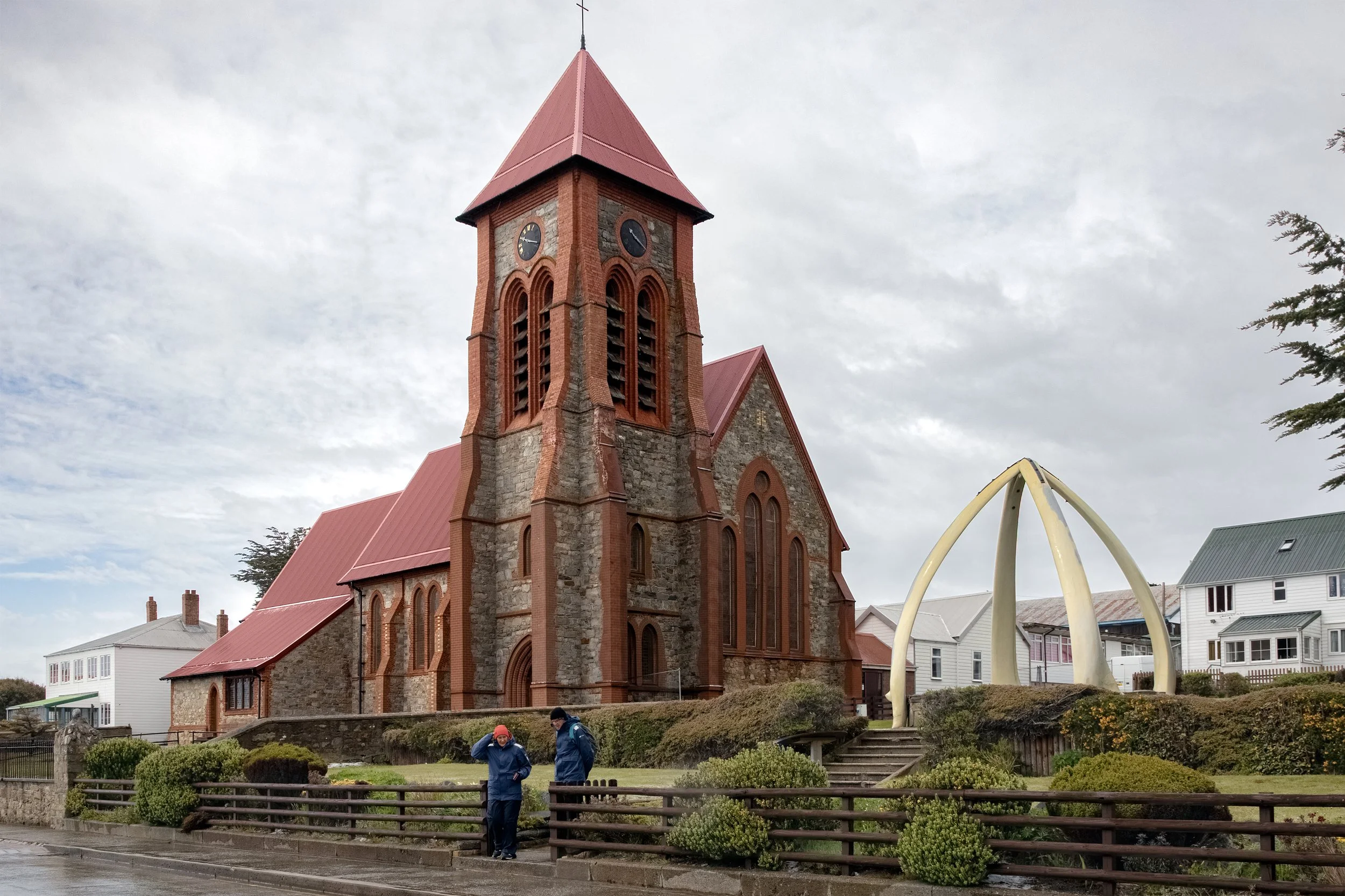 A historic stone and brick church with a tall steeple and clock faces, located in a town with residential homes and a large sculpture in the foreground. Two people with umbrellas are walking along the street.
