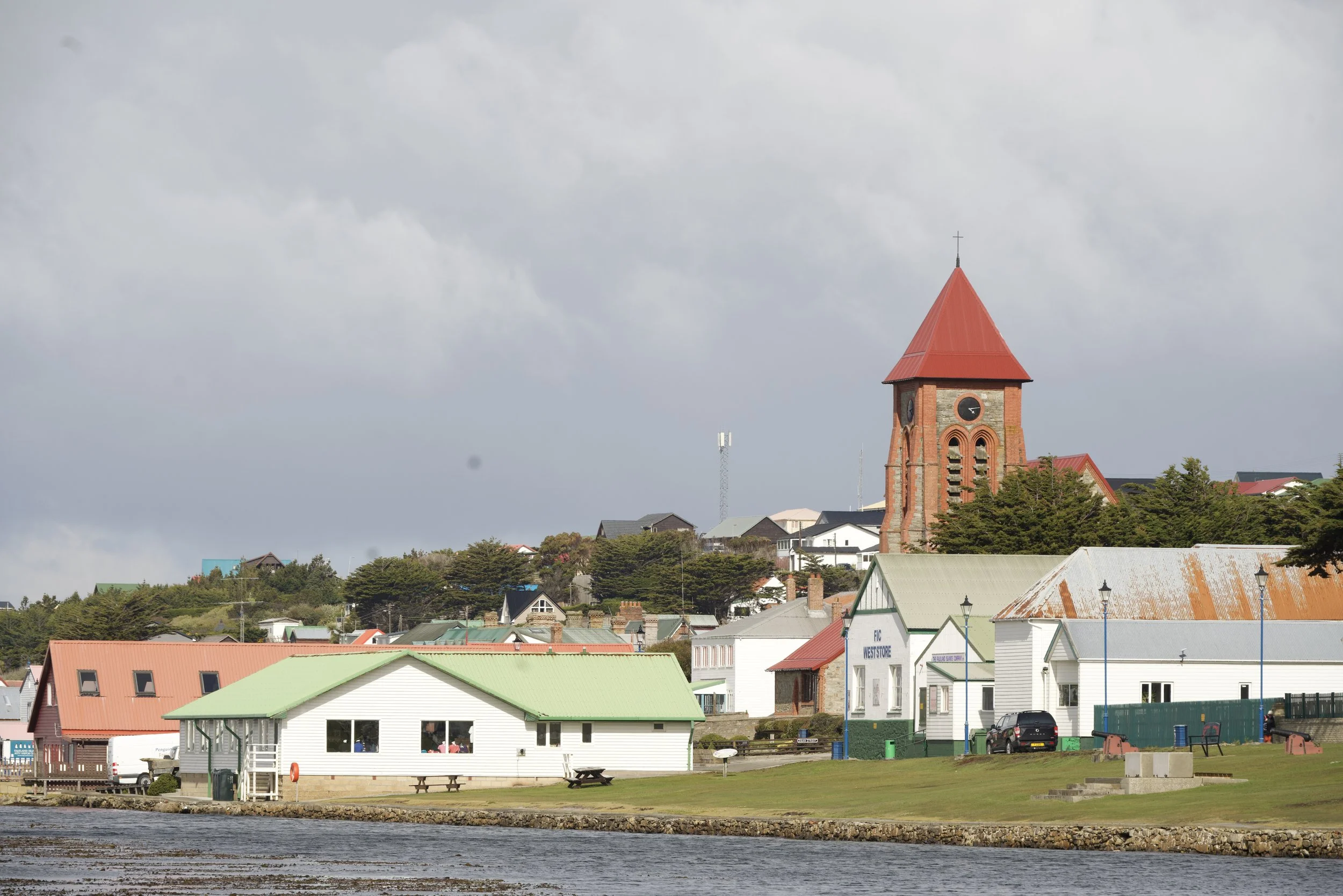 A small town or village with houses and buildings near a body of water, with a prominent church with a red steeple, under a cloudy sky.