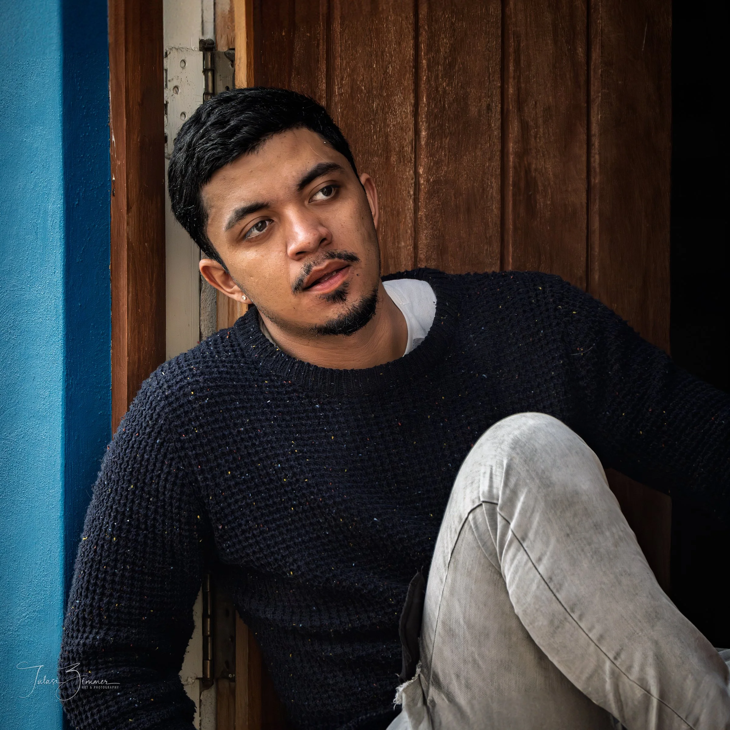 Close-up of a young Mexican man with black hair, mustache, and earrings, lying on a wooden surface with blue background wall, wearing a dark sweater and white pants.
